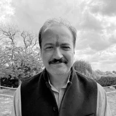 Schwarz-Weiß-Foto eines Mannes mit Bart und Schnurrbart, der draußen steht, im Hintergrund Bäume und Wolken.