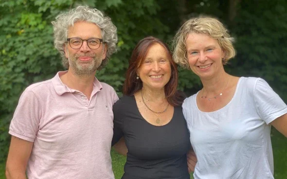 Drei lachende Menschen, zwei Frauen und ein Mann, stehen im Freien vor grünen Bäumen.