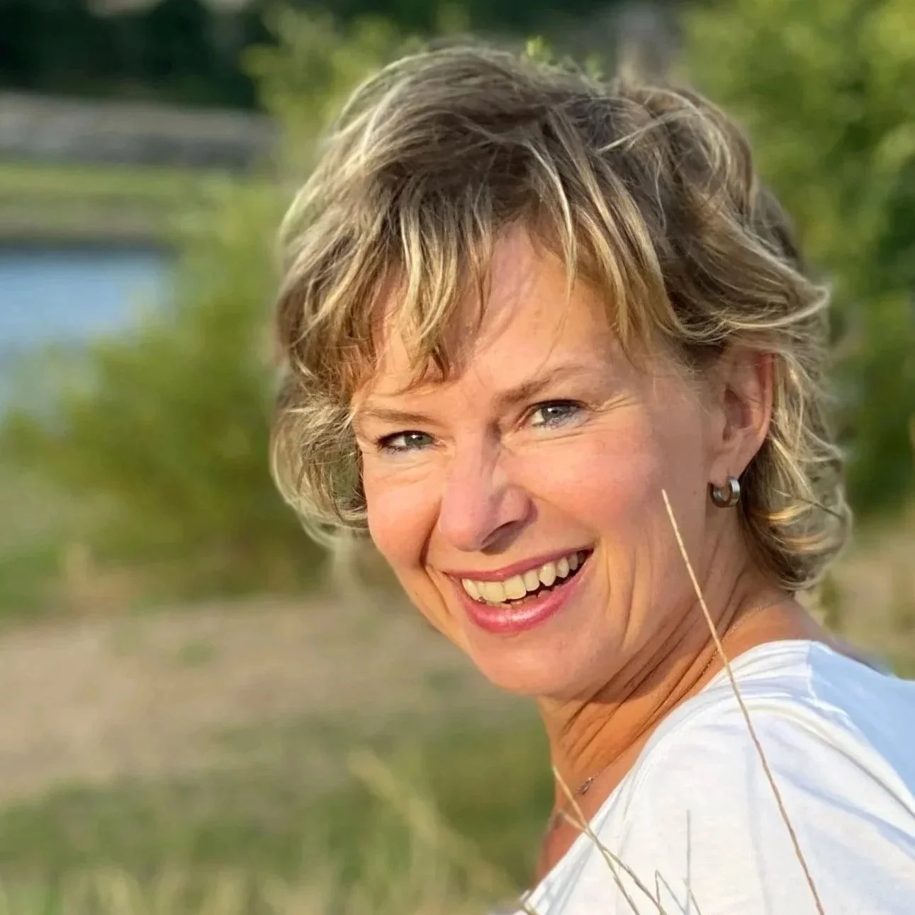 Eine lachende Frau mit kurzen, lockigen Haaren, draußen in der Natur, im Hintergrund Wasser und Bäume.