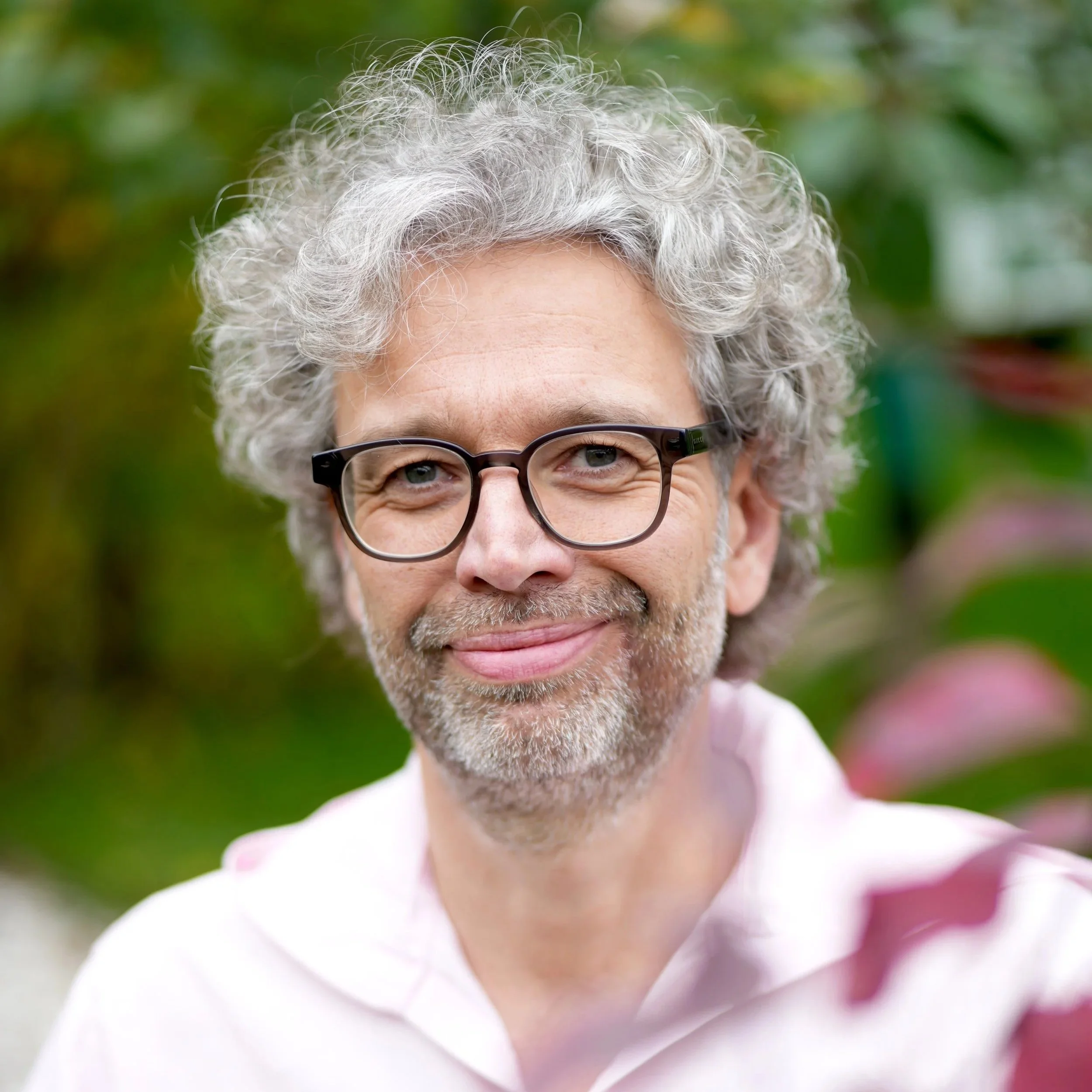 Nahaufnahme eines lächelnden Mannes mit grauem, lockigem Haar, Brille und Bart, im Freien mit grüner Natur im Hintergrund.
