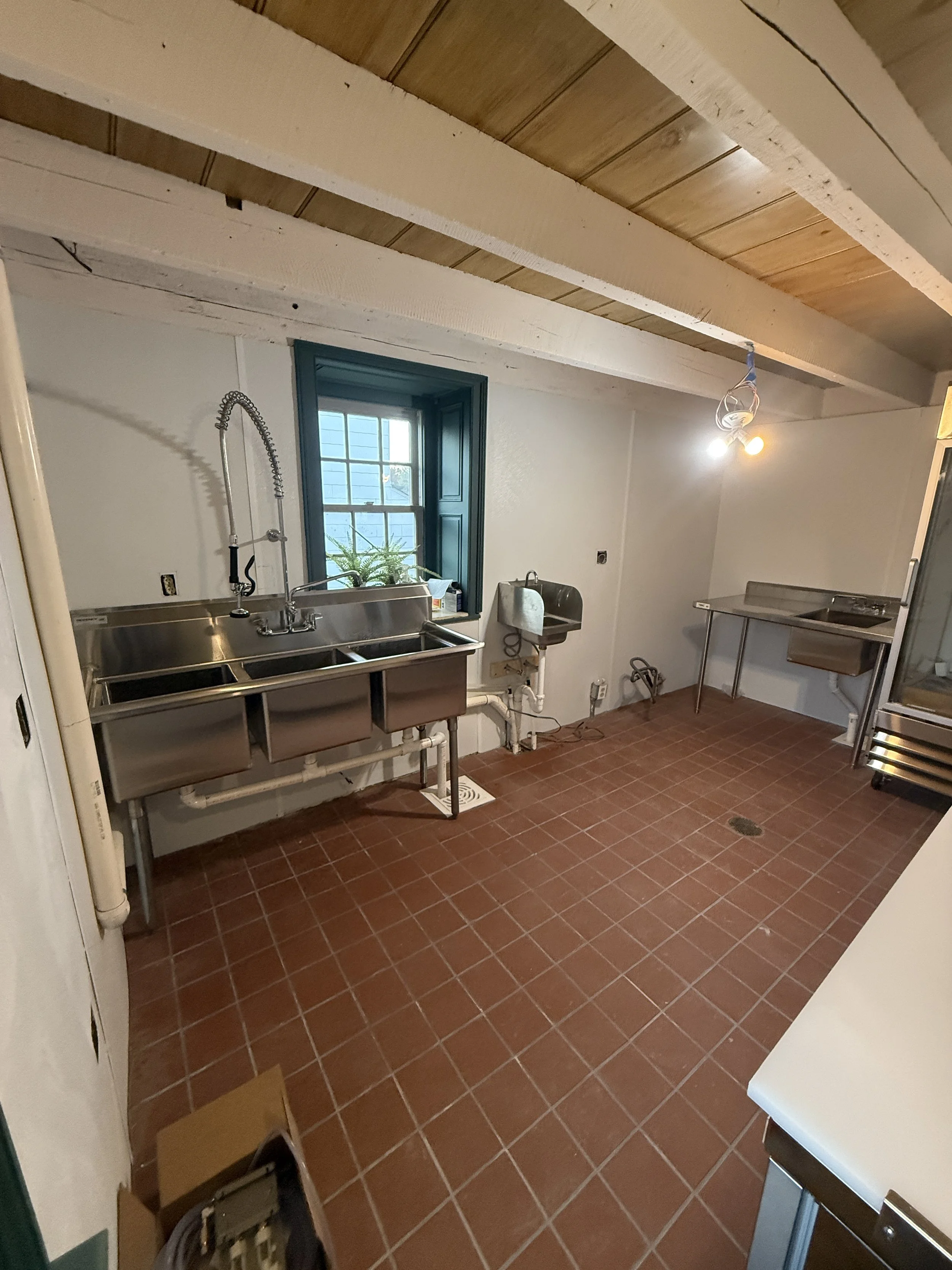 An empty commercial kitchen with a stainless steel double basin sink beneath a window, a small wall-mounted sink, and a stainless steel table. The room has white walls, exposed wooden beams on the ceiling, terracotta floor tiles, and a single lightbulb hanging from the ceiling.