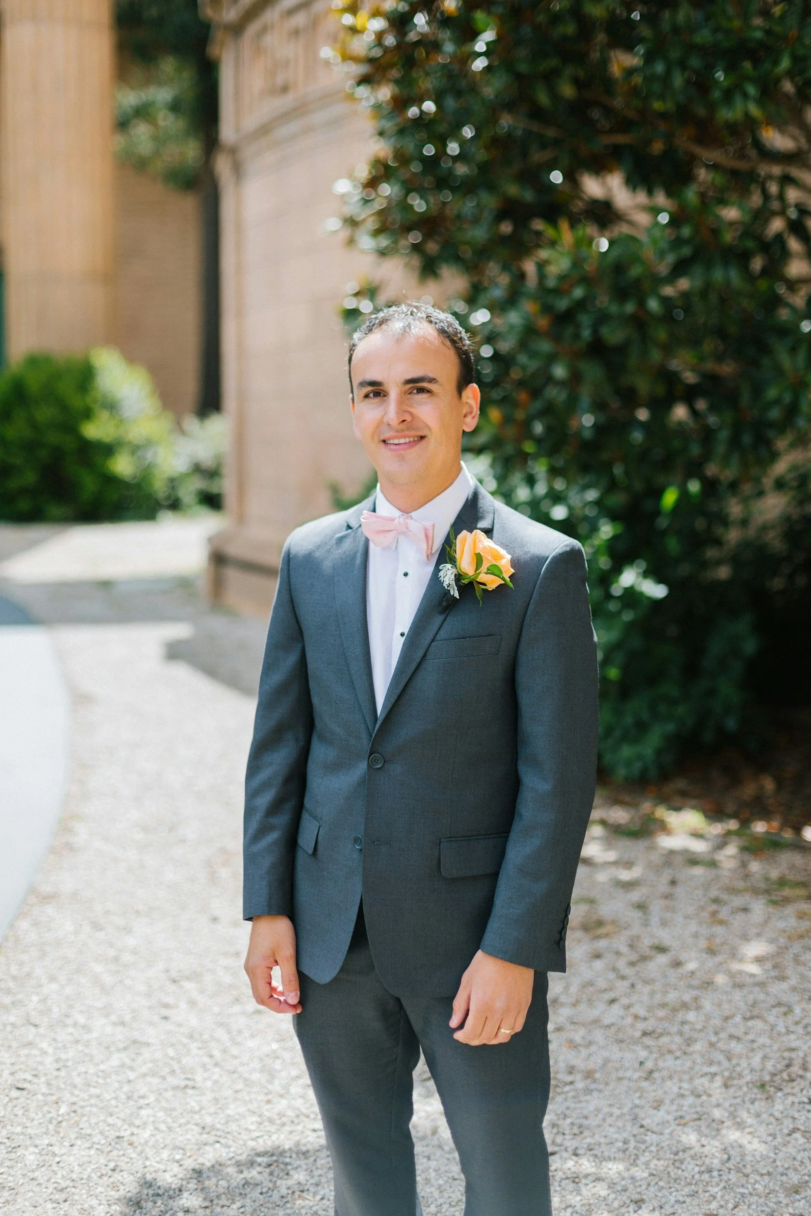A man in a gray suit with a pink bow tie and a yellow rose boutonniere standing outdoors on a sunny day, smiling.