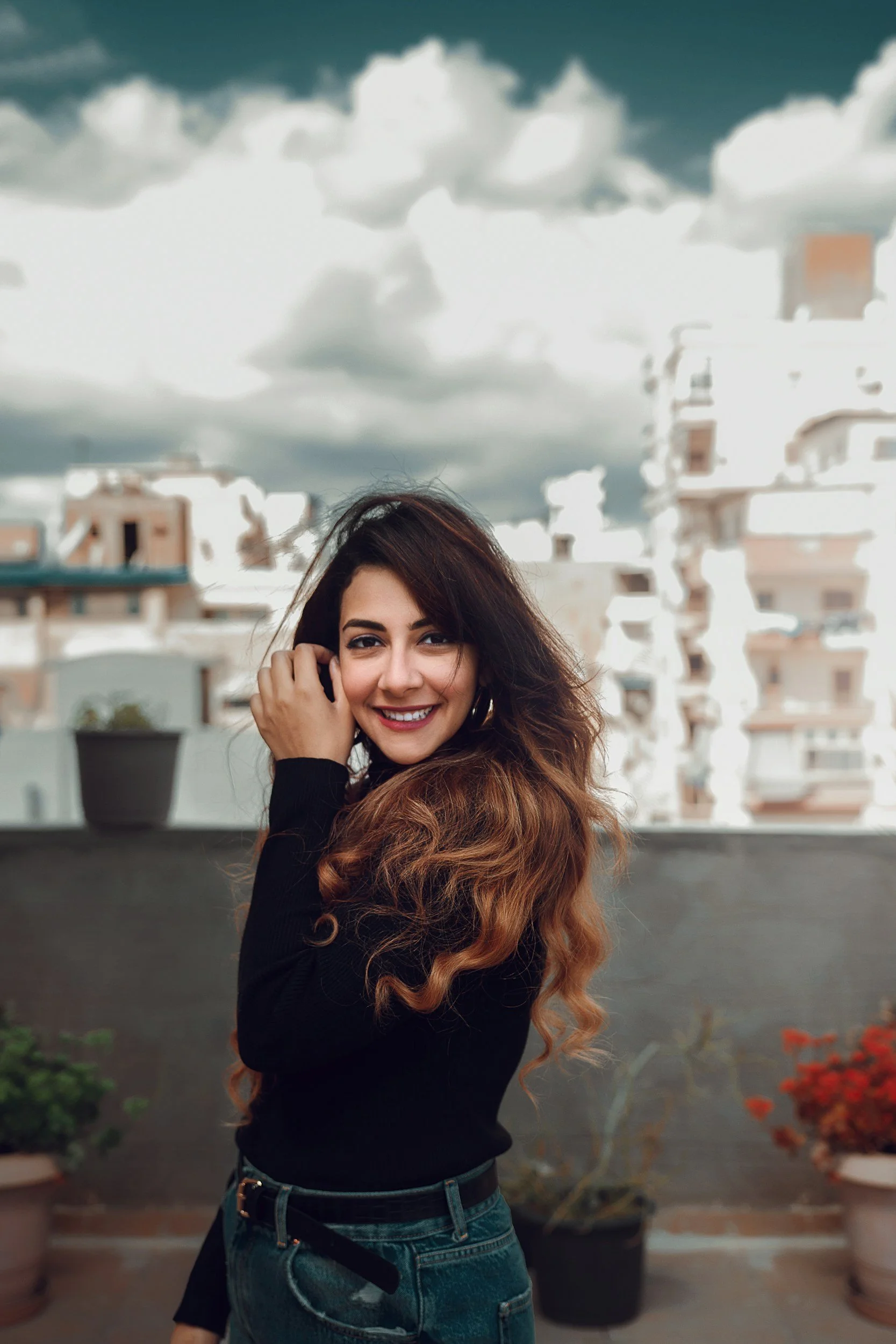 Smiling woman with long, wavy brown hair standing outdoors on a balcony with potted plants and city buildings in the background, under a cloudy sky.