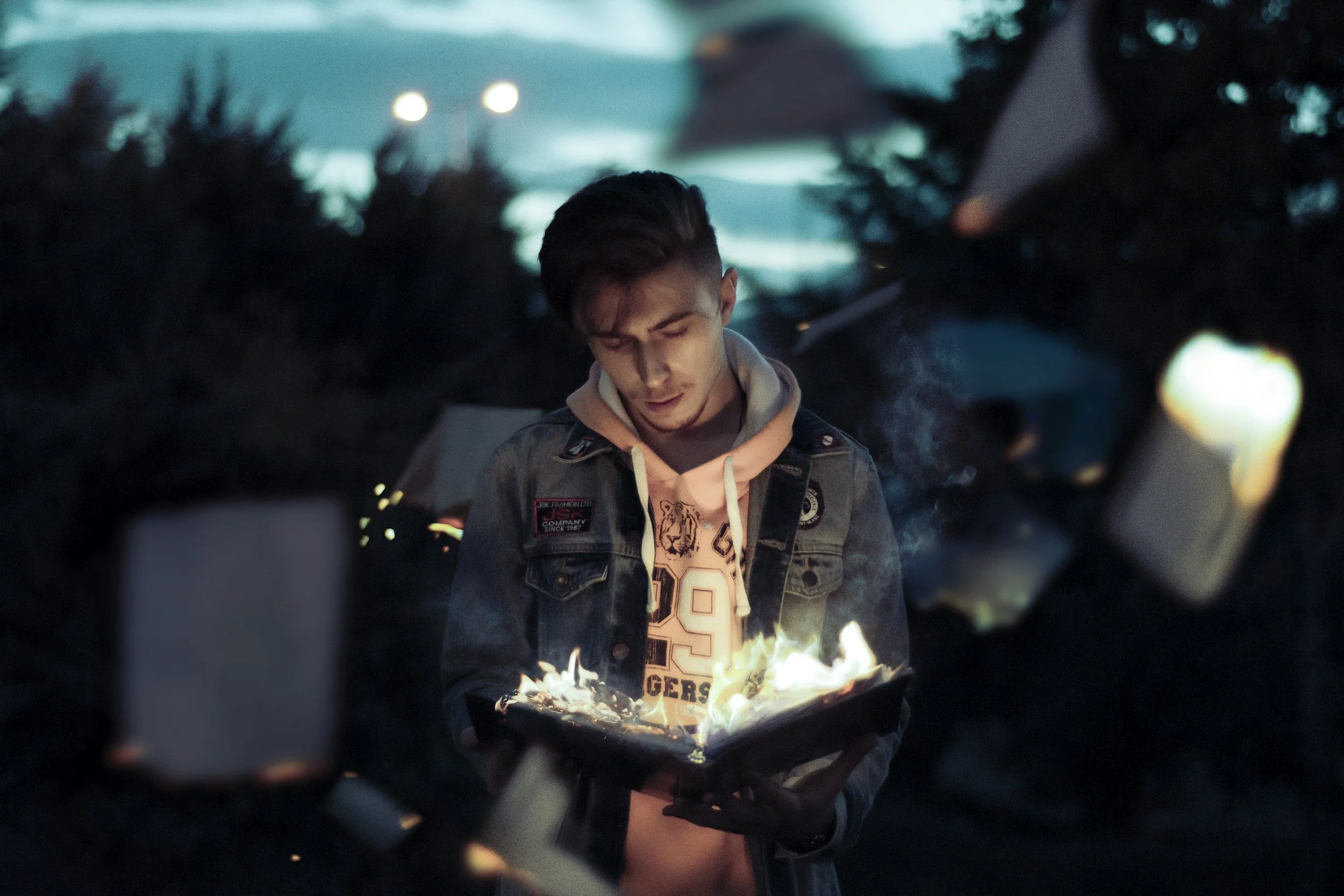 A young man stands outdoors at dusk or evening, holding a book with flames and smoke coming from it, with a focused and concerned expression.