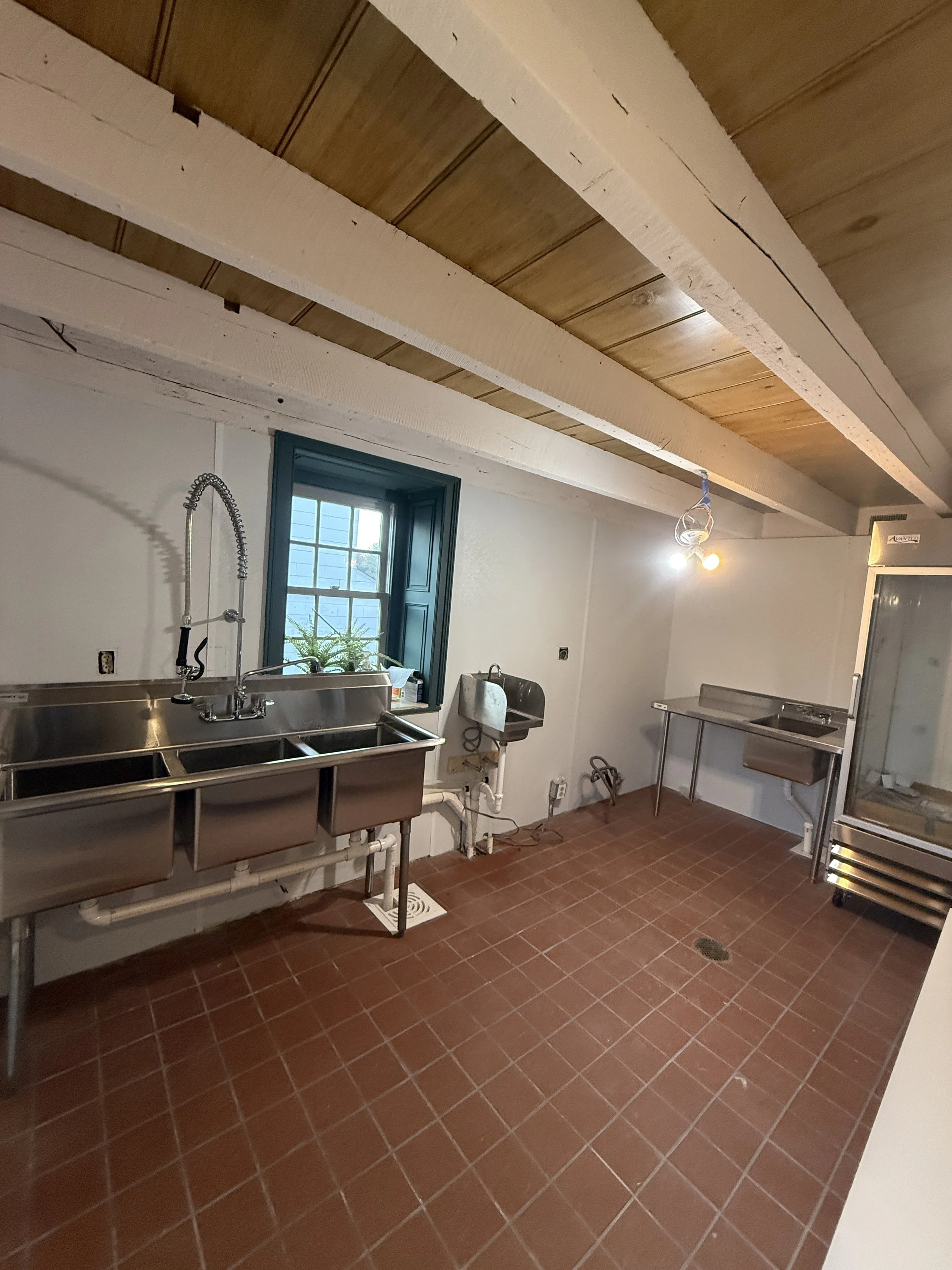 Empty commercial kitchen with red tile floor, stainless steel sink, small window, and three working lights hanging from the ceiling with exposed wooden beams.