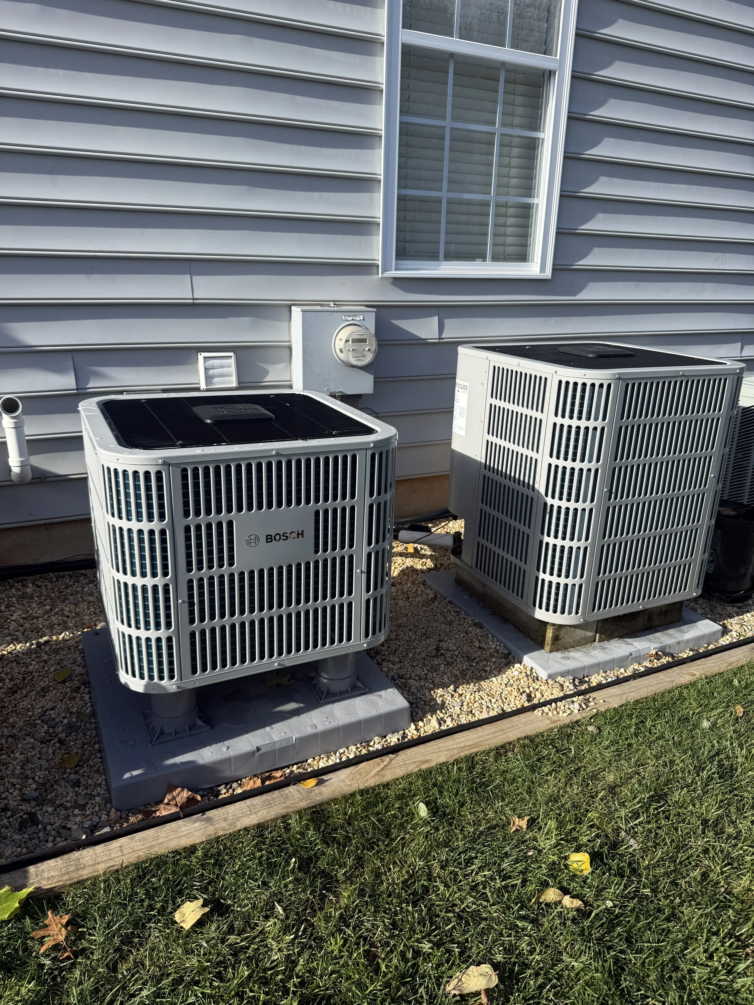 Two outdoor air conditioning units next to a house with gray siding and a window.