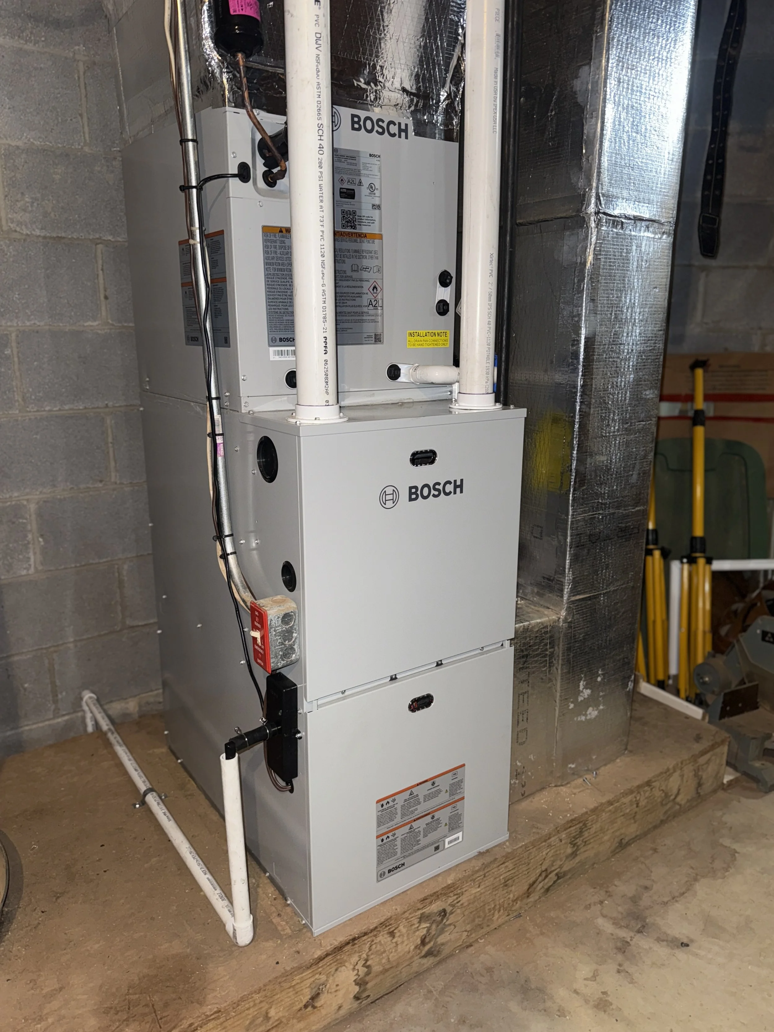 A Bosch HVAC unit installed on a concrete floor, with pipes and wiring connected to it, situated in a basement or utility room with cinder block walls and insulation-covered ductwork nearby.