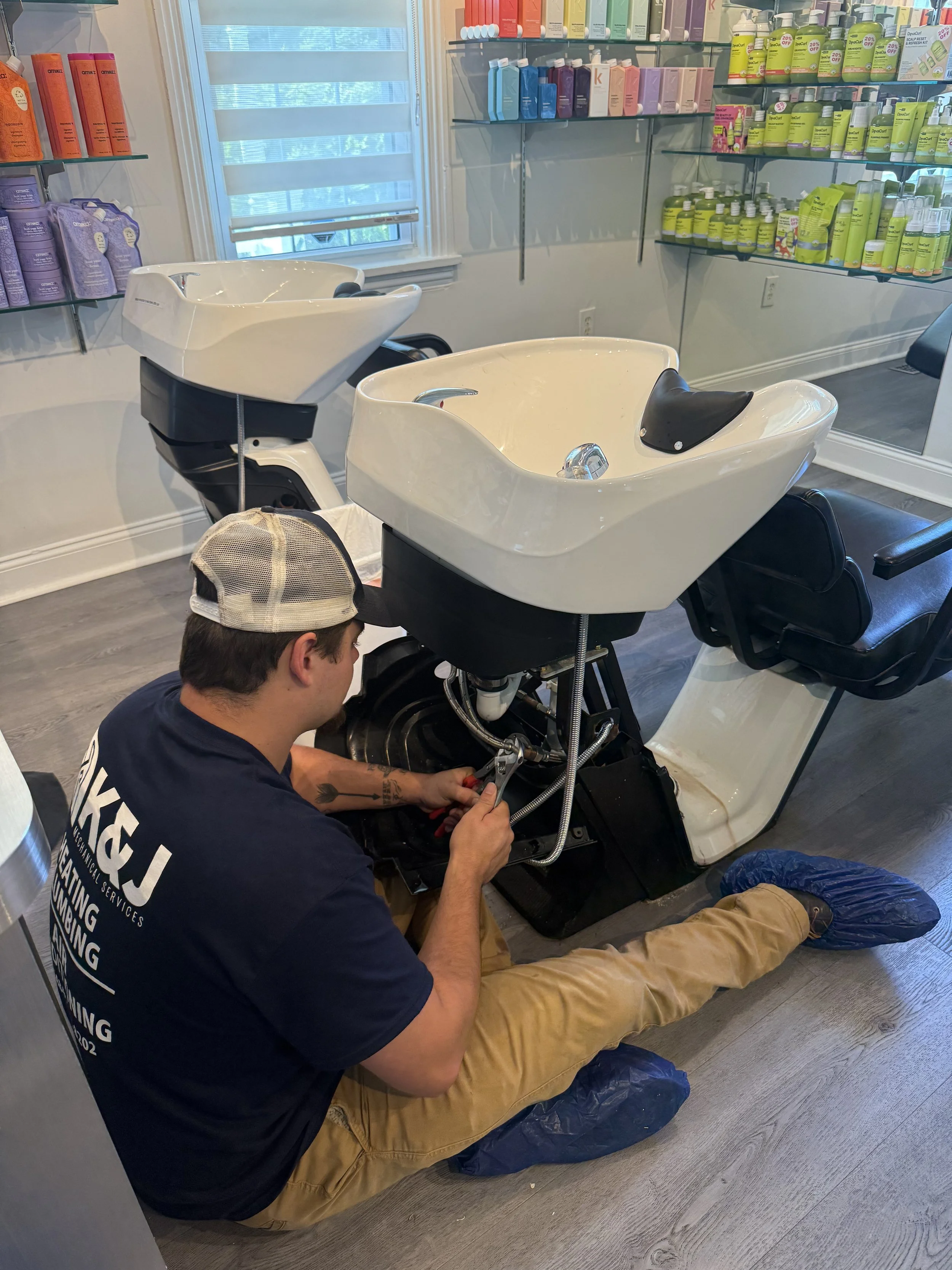 A man wearing a cap and a t-shirt, sitting on the floor, repairing a salon hair washing station in a salon. The background shows shelves with hair care products and a window with blinds.