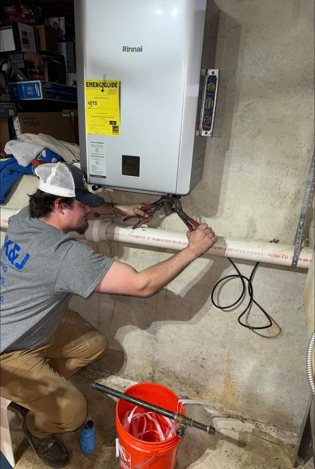 A man installing or repairing a water heater in a basement, using pipe wrenches. There is a large white water heater labeled 'Rinnai' and a bucket with plumbing tools.