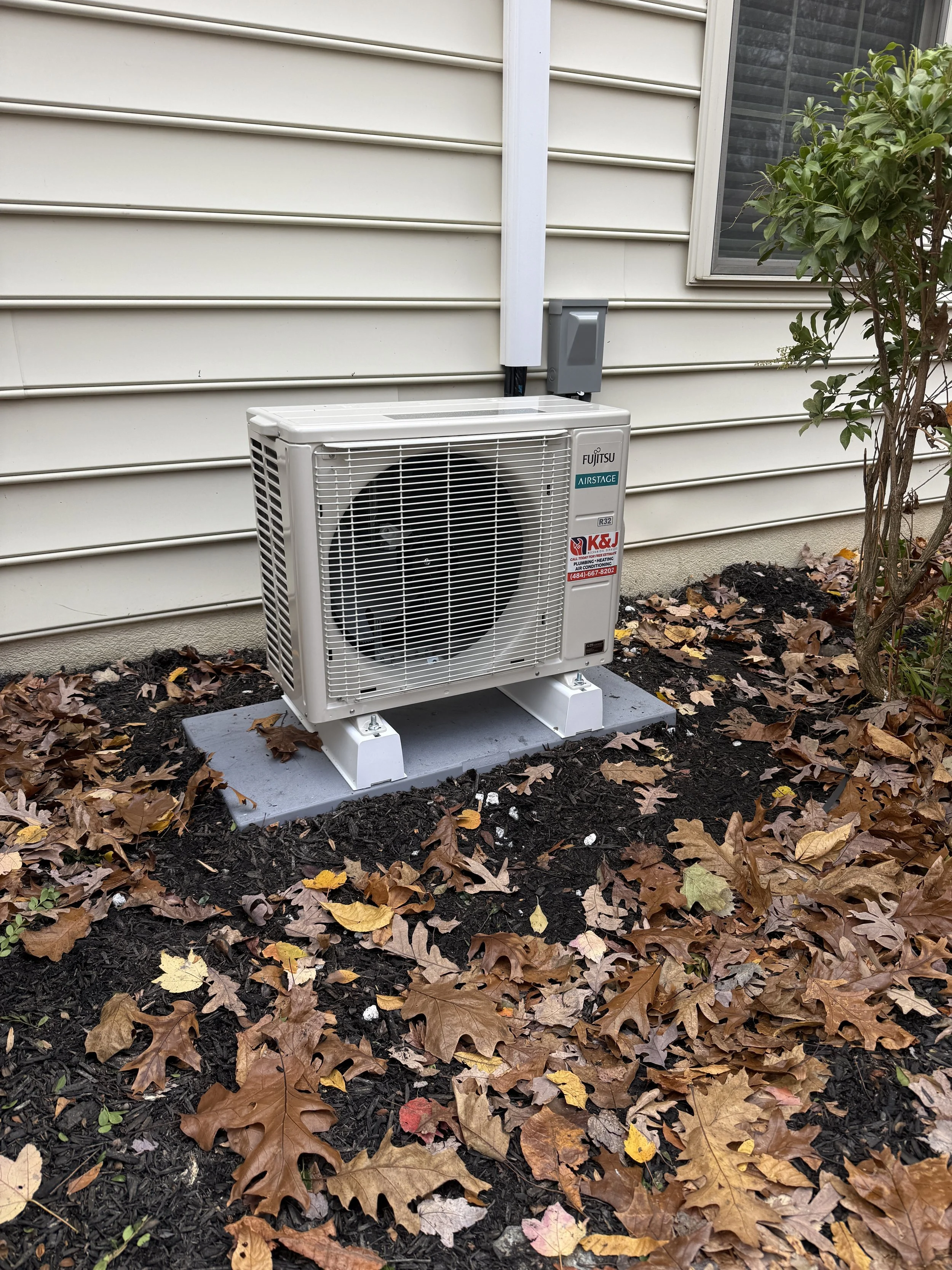 Fujitsu air conditioning outdoor unit installed next to a house with beige siding, on a concrete pad, surrounded by fallen autumn leaves and a small bush.