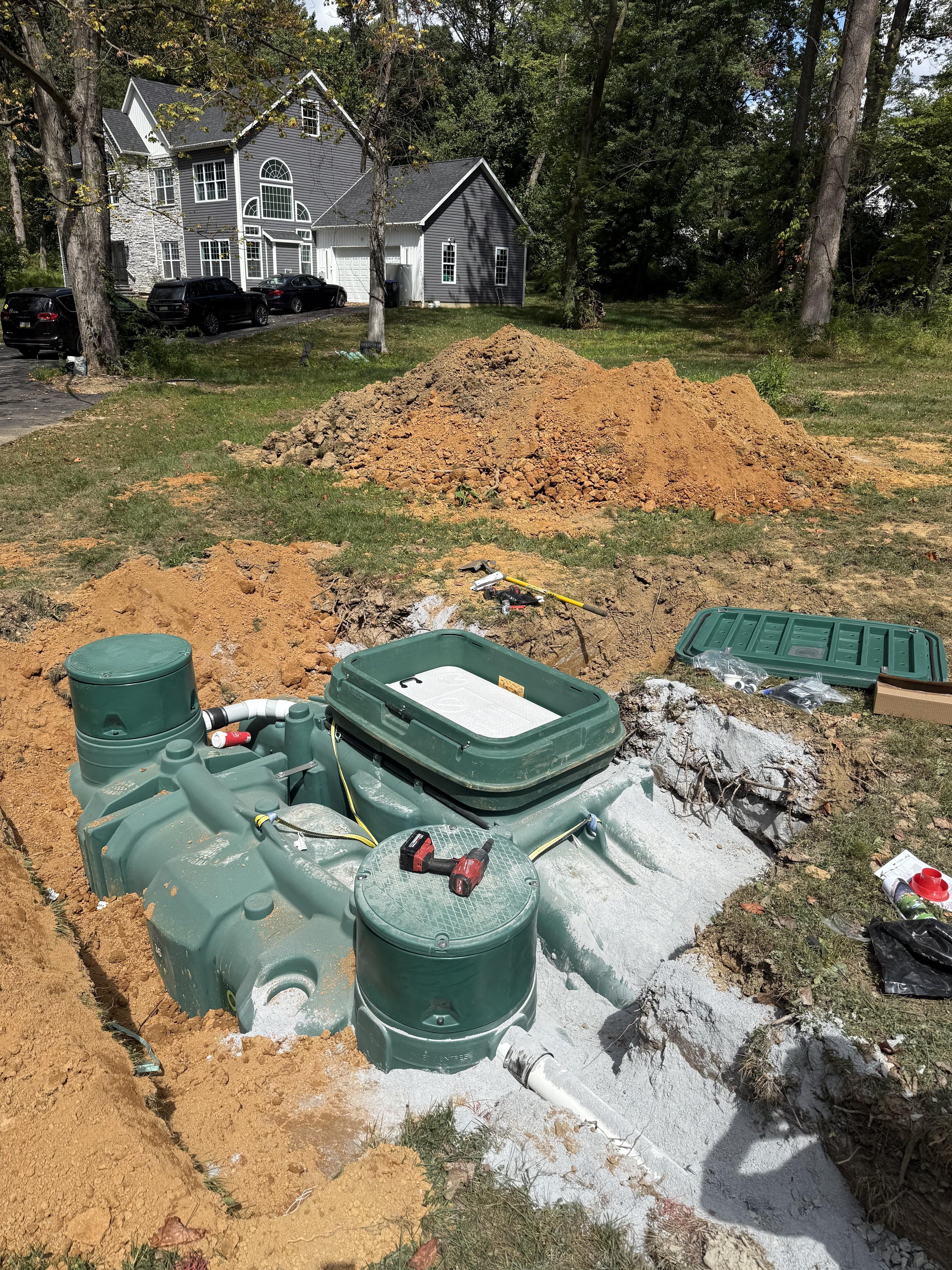 Installation of a new septic system in a backyard, with a green septic tank partly buried in a hole, surrounded by piles of dirt and construction tools, with a house and trees in the background.
