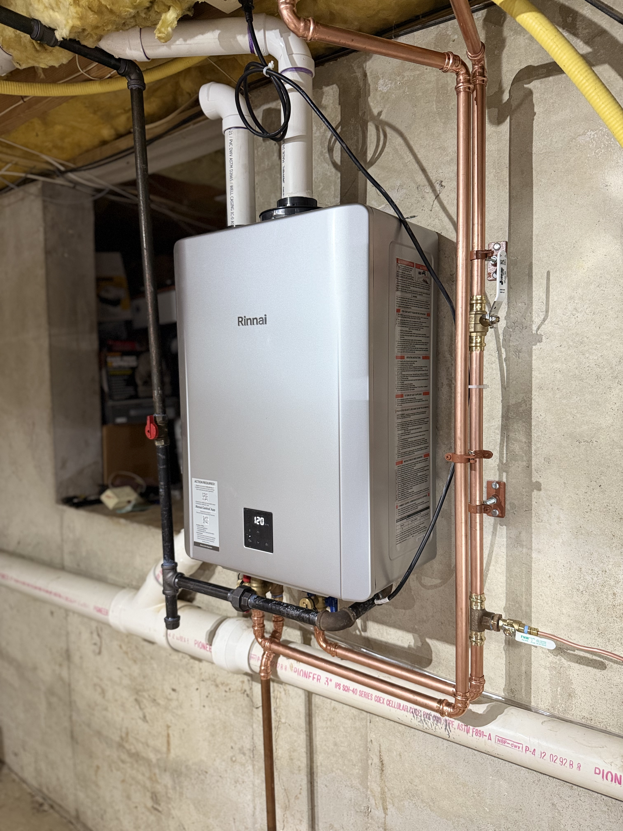 A Rinnai tankless water heater mounted on a concrete wall, with copper and PVC pipes connected to it, located in a basement or utility room.