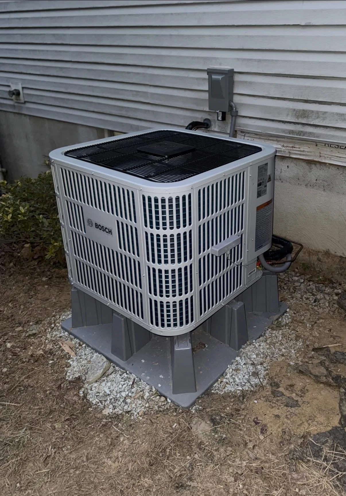 Outside view of a Bosch air conditioning or heat pump unit installed on a raised platform next to a house wall.