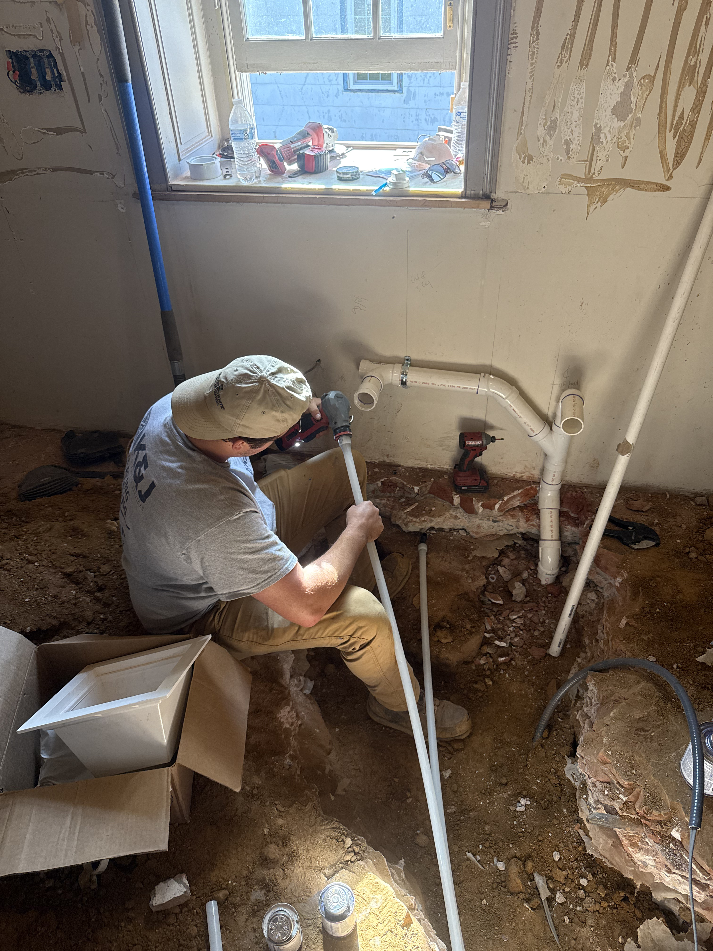 A worker installing plumbing pipes in a basement or crawl space, with exposed dirt and construction tools around.