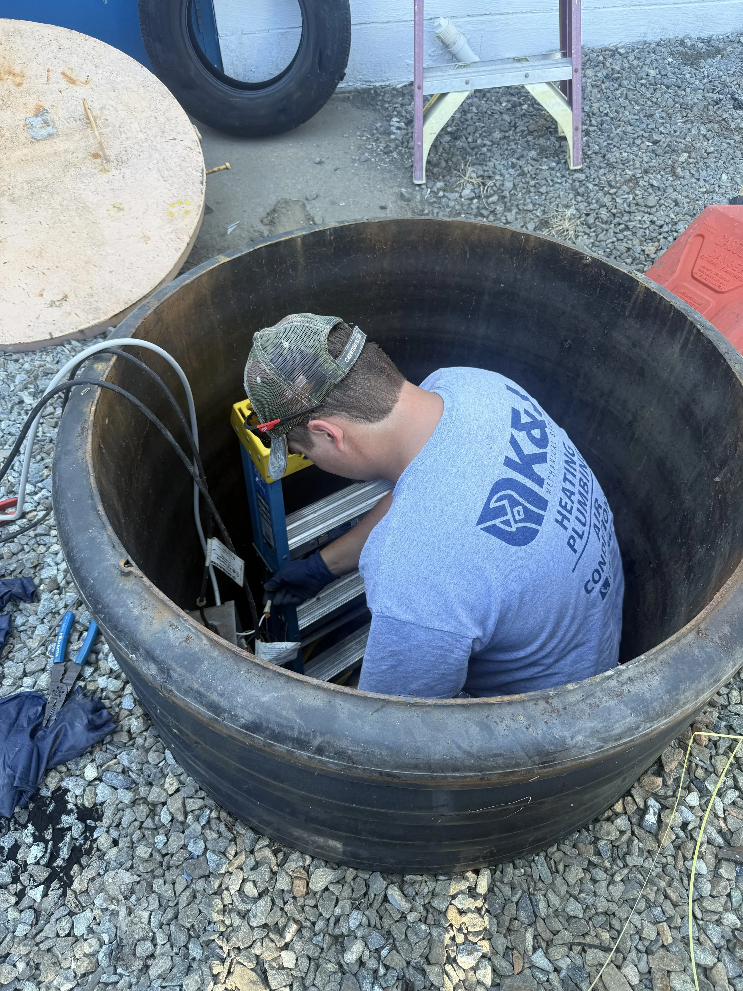 A man working inside a large underground sewer pit, installing a new pump. Repairing electrical cables with tools nearby.