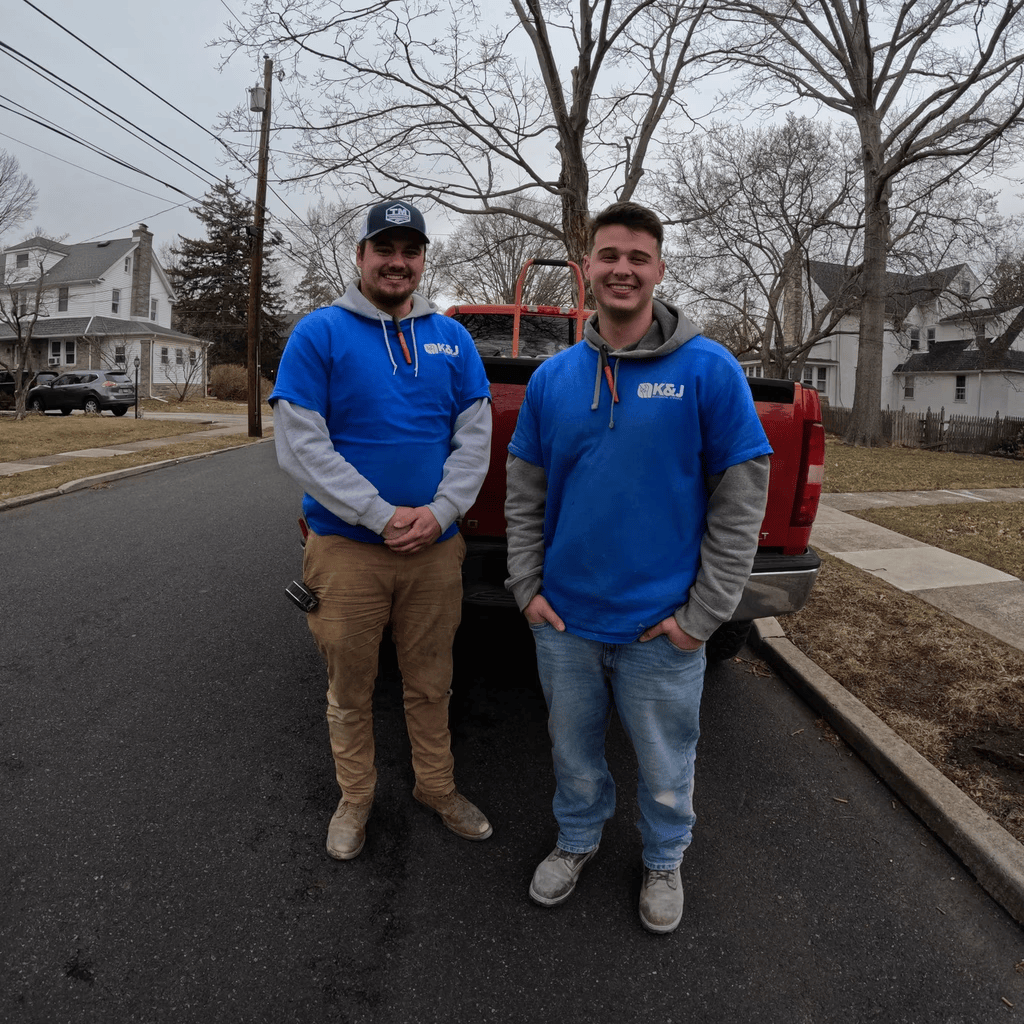 Two men wearing blue K&J shirts stand outdoors on a street in front of a red pickup truck, with residential houses and leafless trees in the background, on a cloudy day.