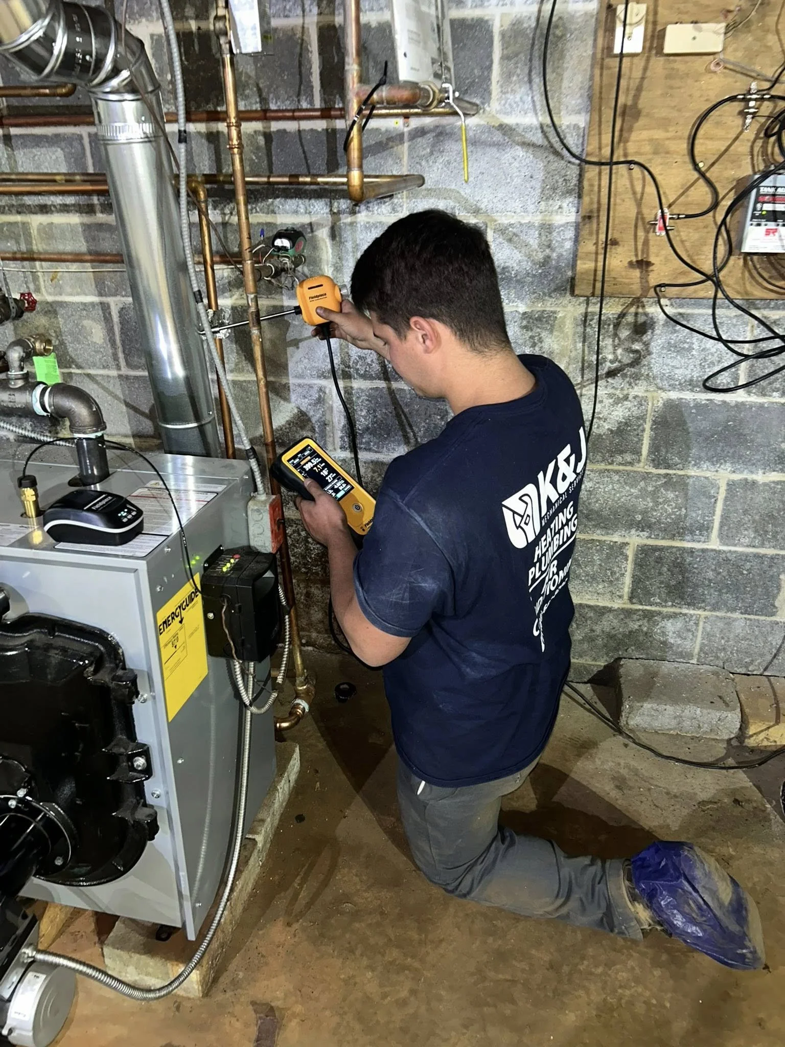 A technician kneels on the floor while using a multimeter to test plumbing pipes in an unfinished basement with concrete block walls.