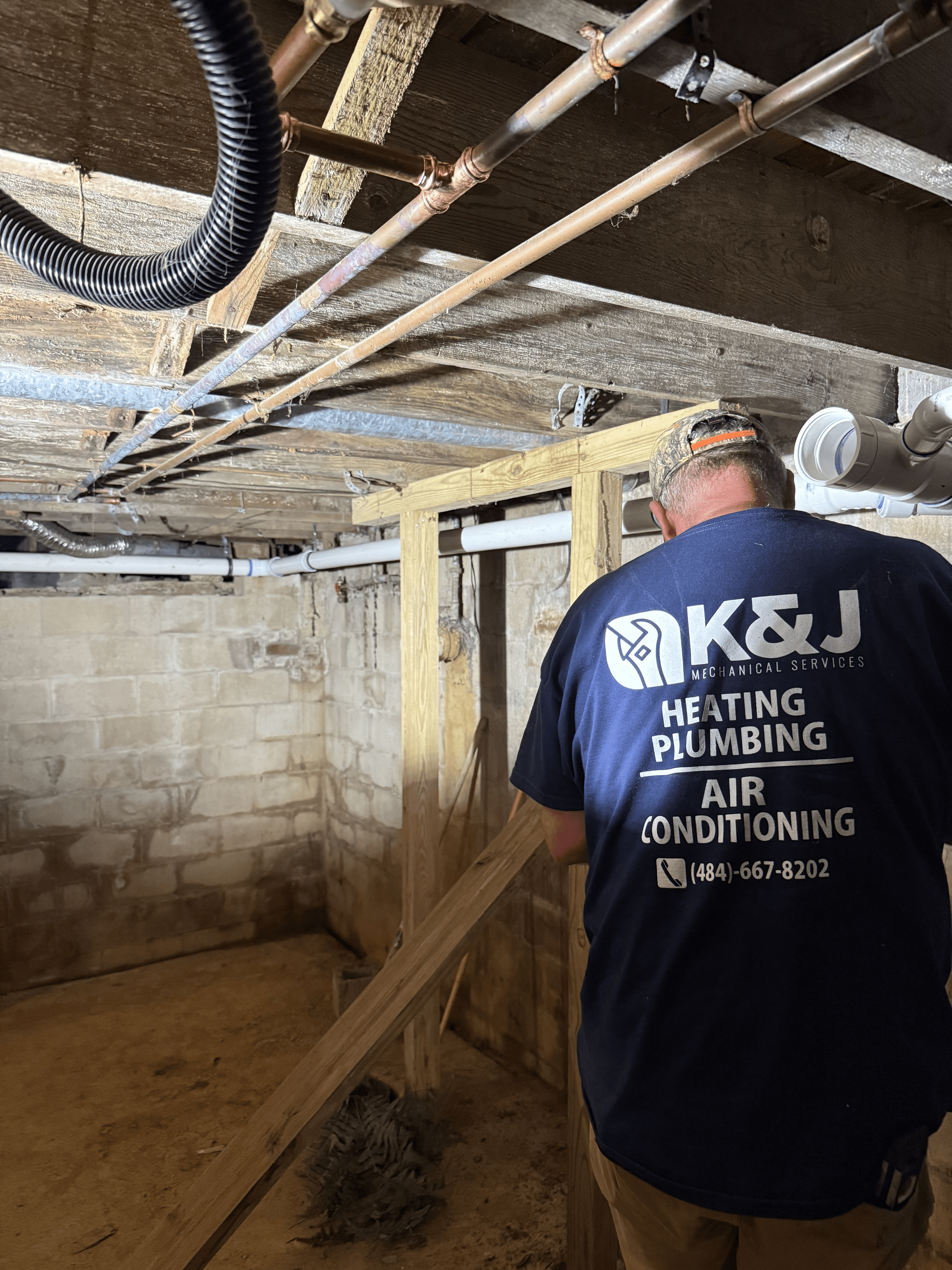 A worker wearing a K&J Mechanical Services t-shirt is inspecting and working on plumbing and HVAC systems in a building under construction or renovation.