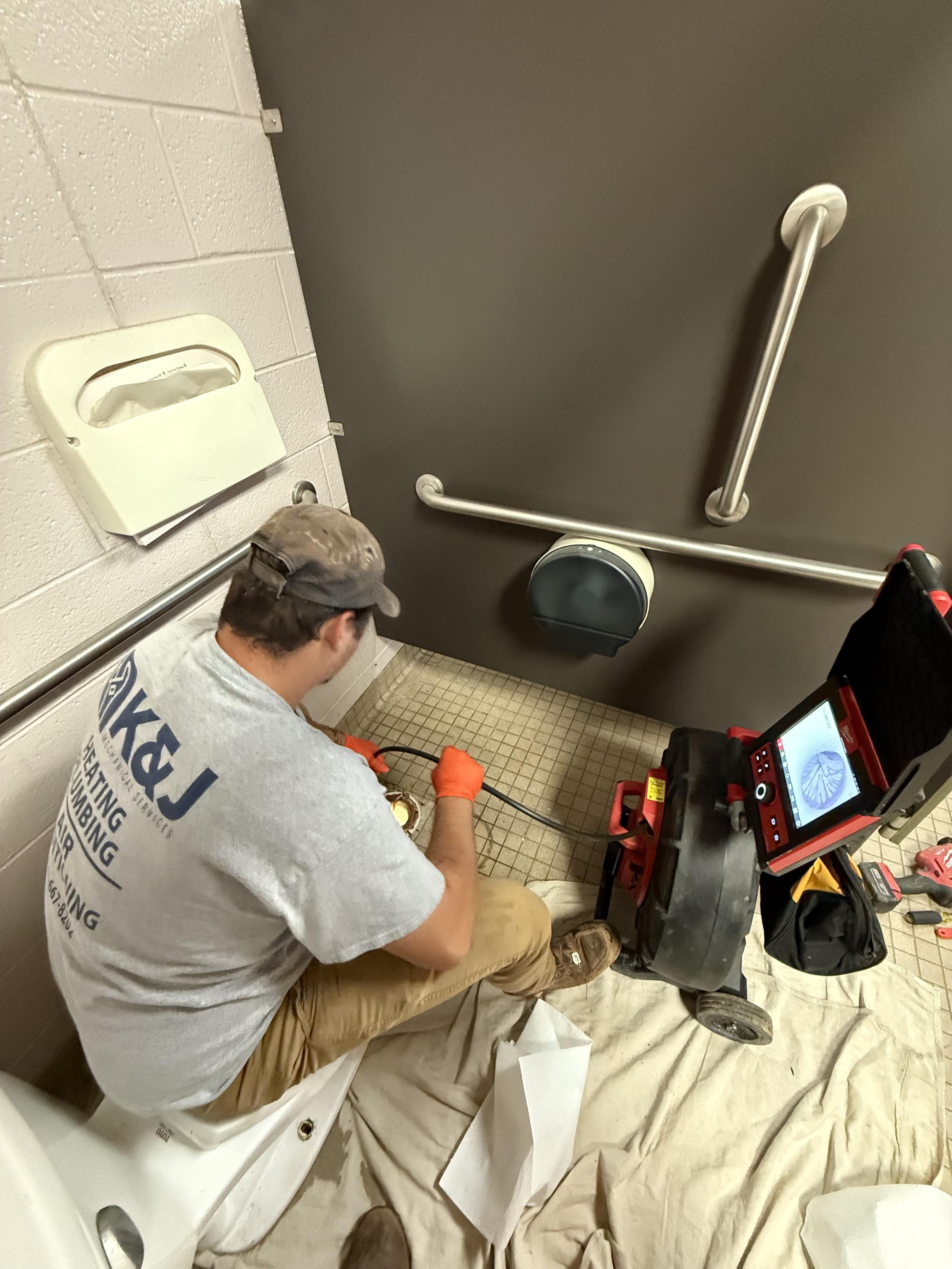 A plumber performing maintenance or repairs inside a commercial or public restroom. The plumber is seated on the toilet, wearing a gray T-shirt, khaki pants, and orange gloves, using tools. A drain cleaning machine is positioned on a beige cloth on t