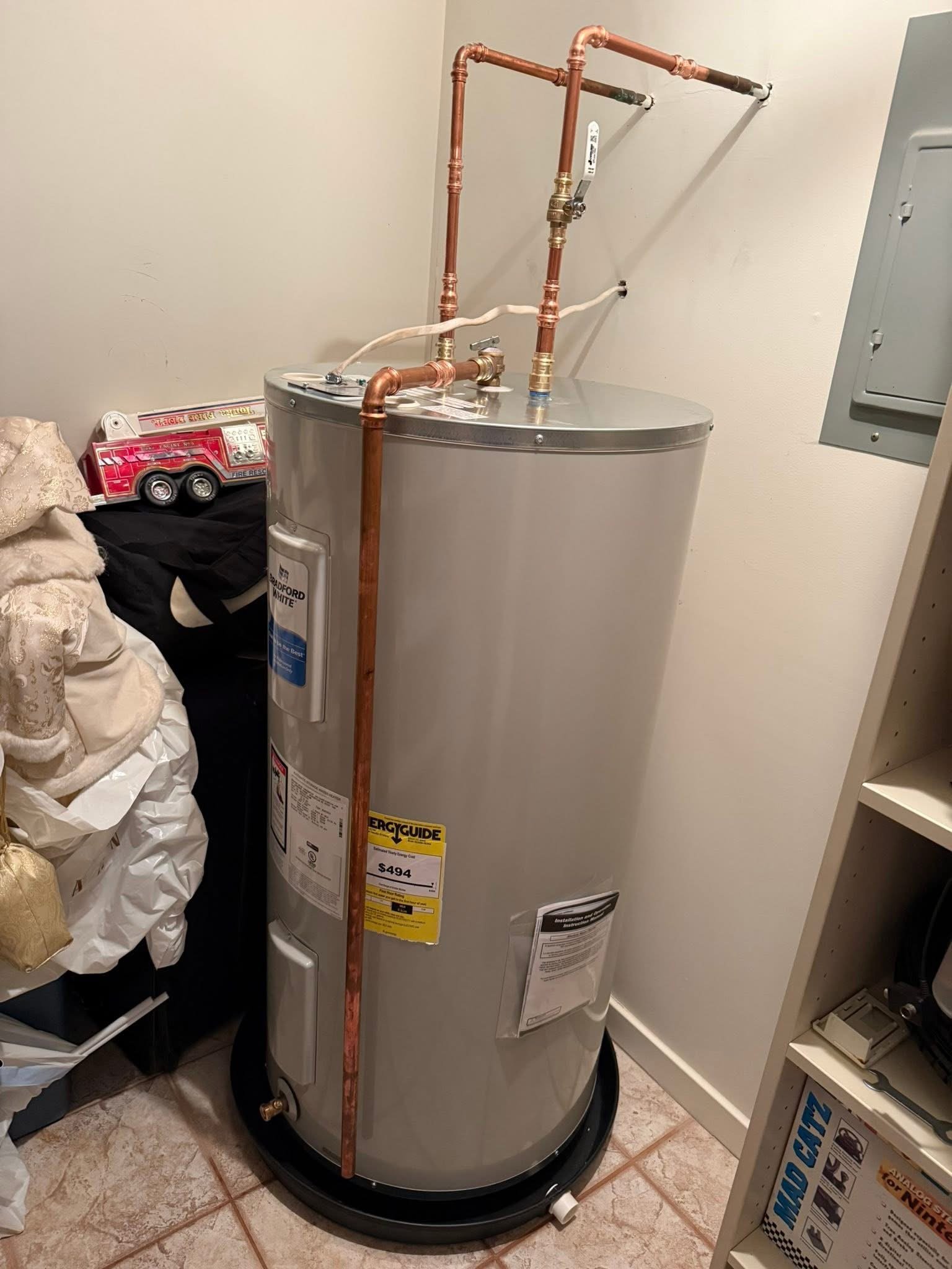 A water heater with copper pipes installed on top, situated in a corner closet with a white wall and a gray utility box, surrounded by shelves and miscellaneous items.