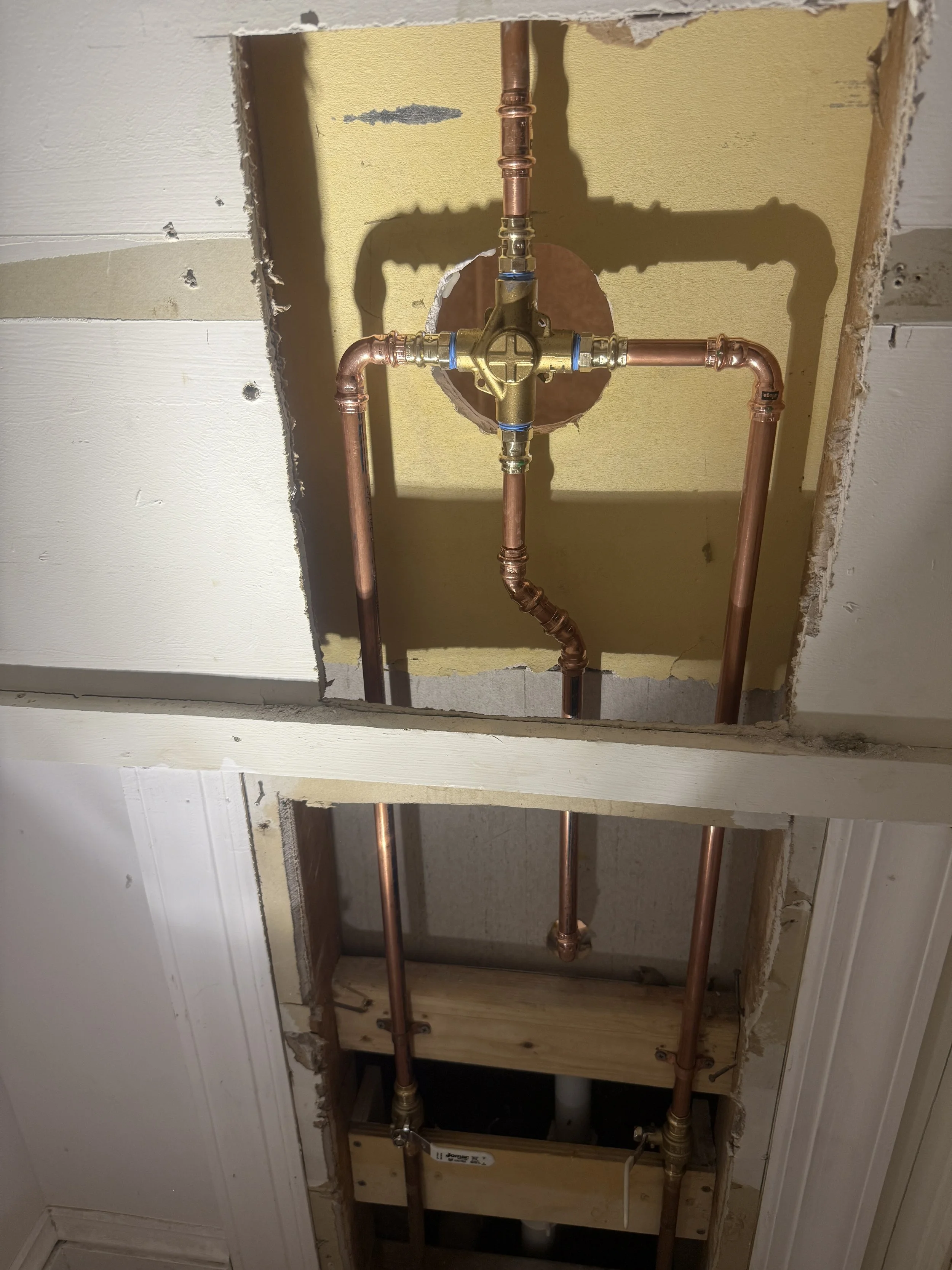 Copper water supply plumbing setup in a wall, with pipes and fittings, including a central valve and a cut-out section of wall revealing the plumbing system.