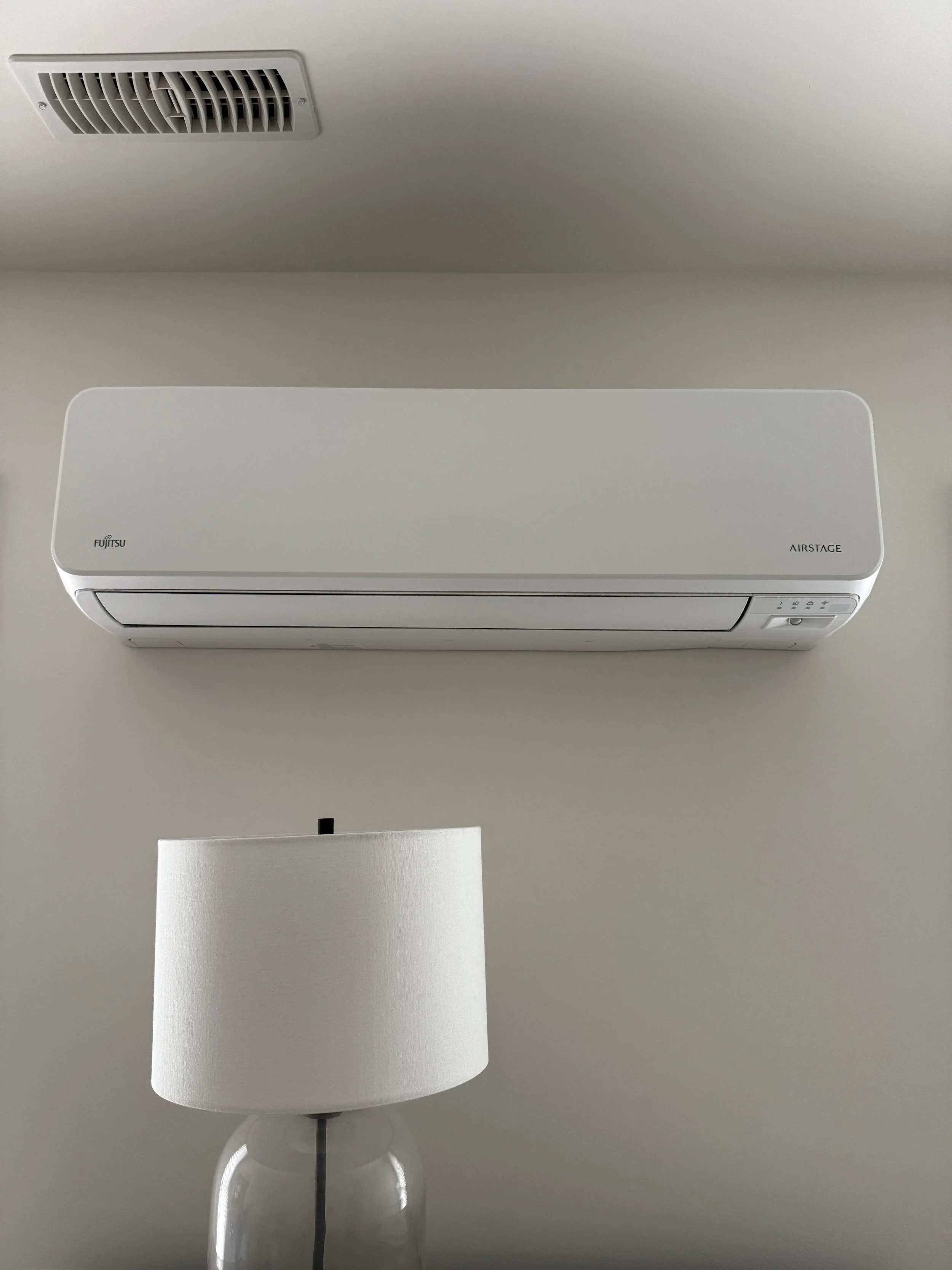 Wall-mounted Fujitsu air conditioner unit above a white lampshade on a glass base.