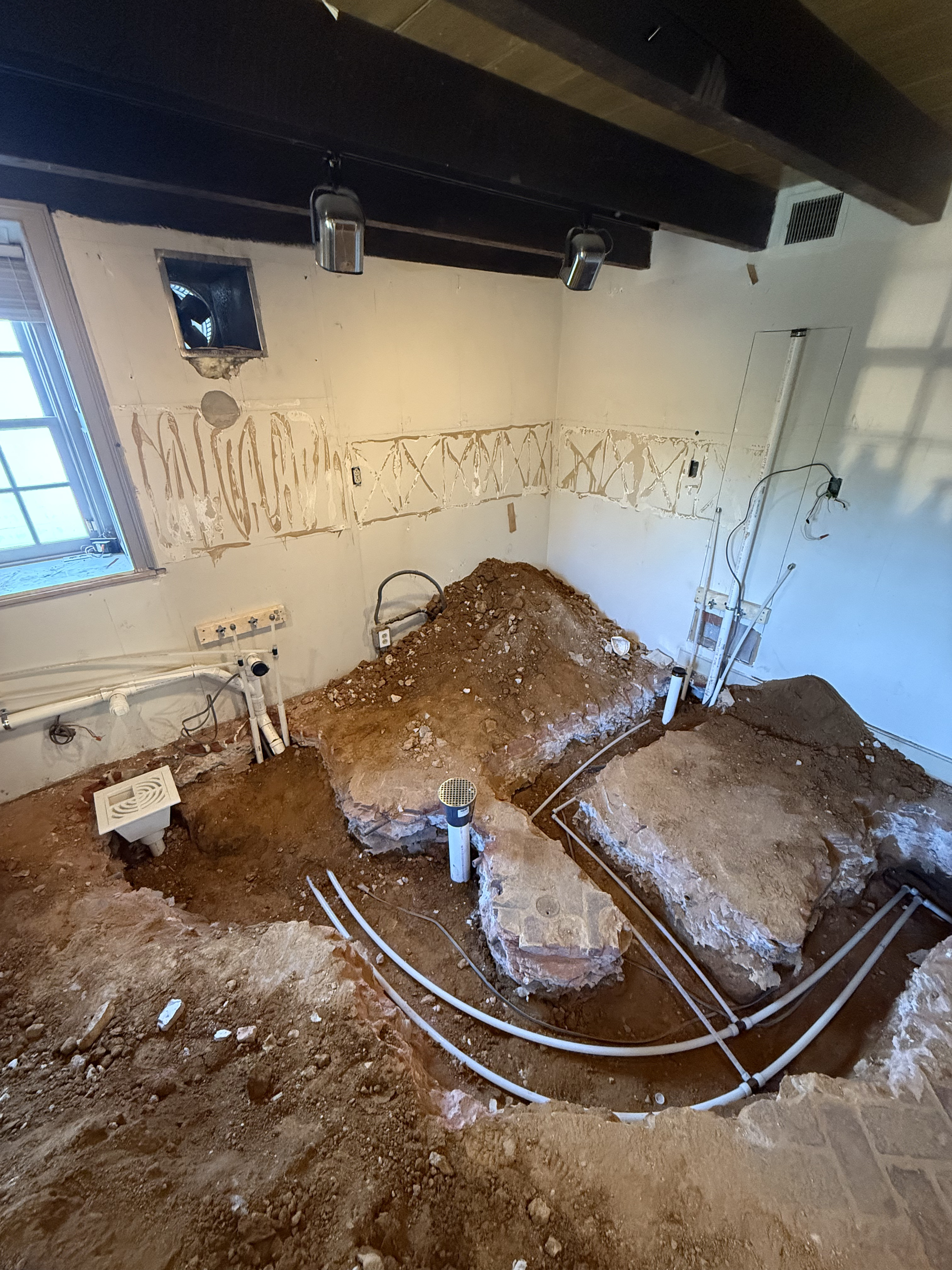 A room under renovation with exposed pipes, plumbing, and dirt on the floor, and a large dug-out area in the center.