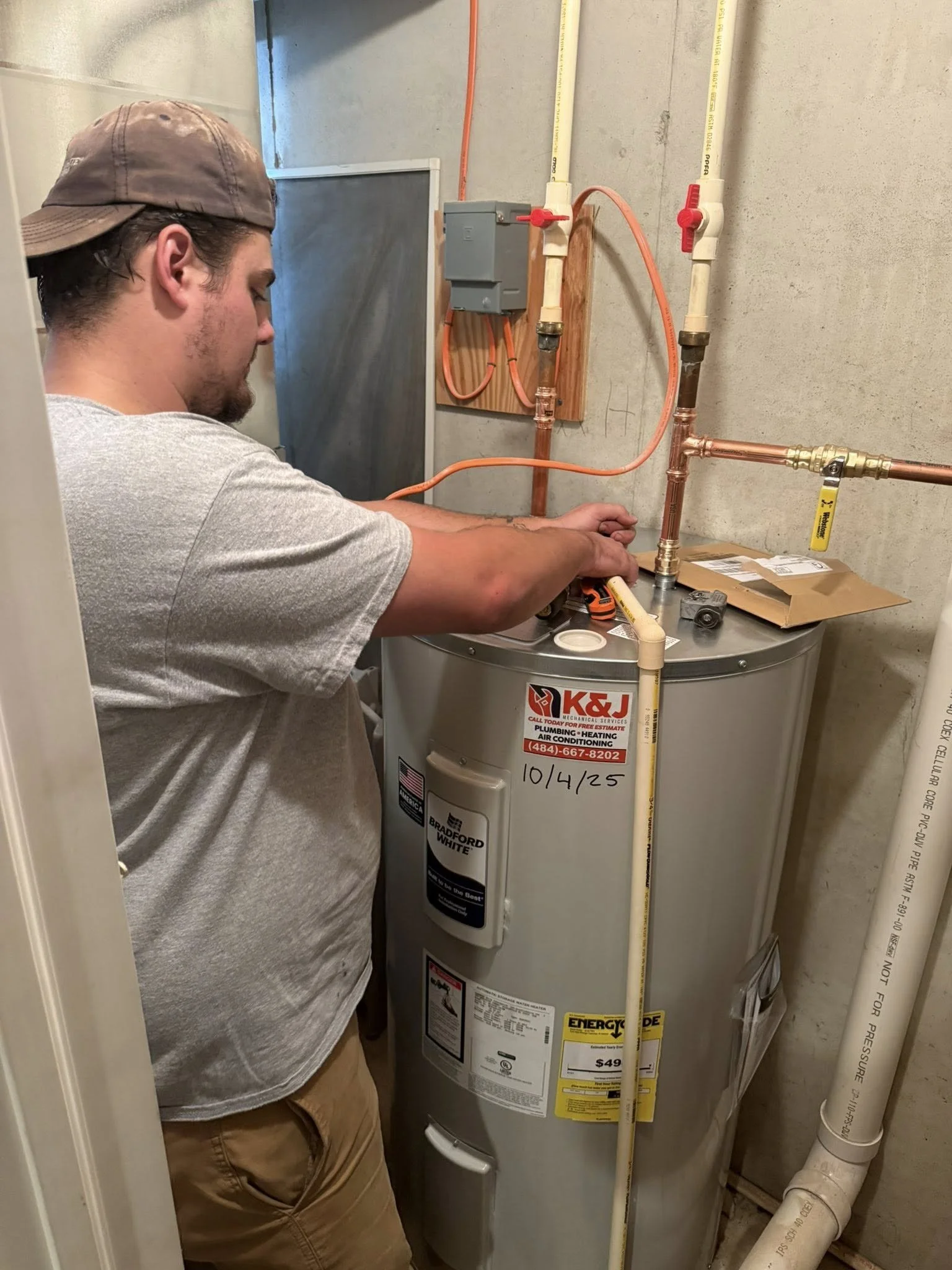 A man working on a water heater in a utility closet, with copper pipes and electrical wiring visible.