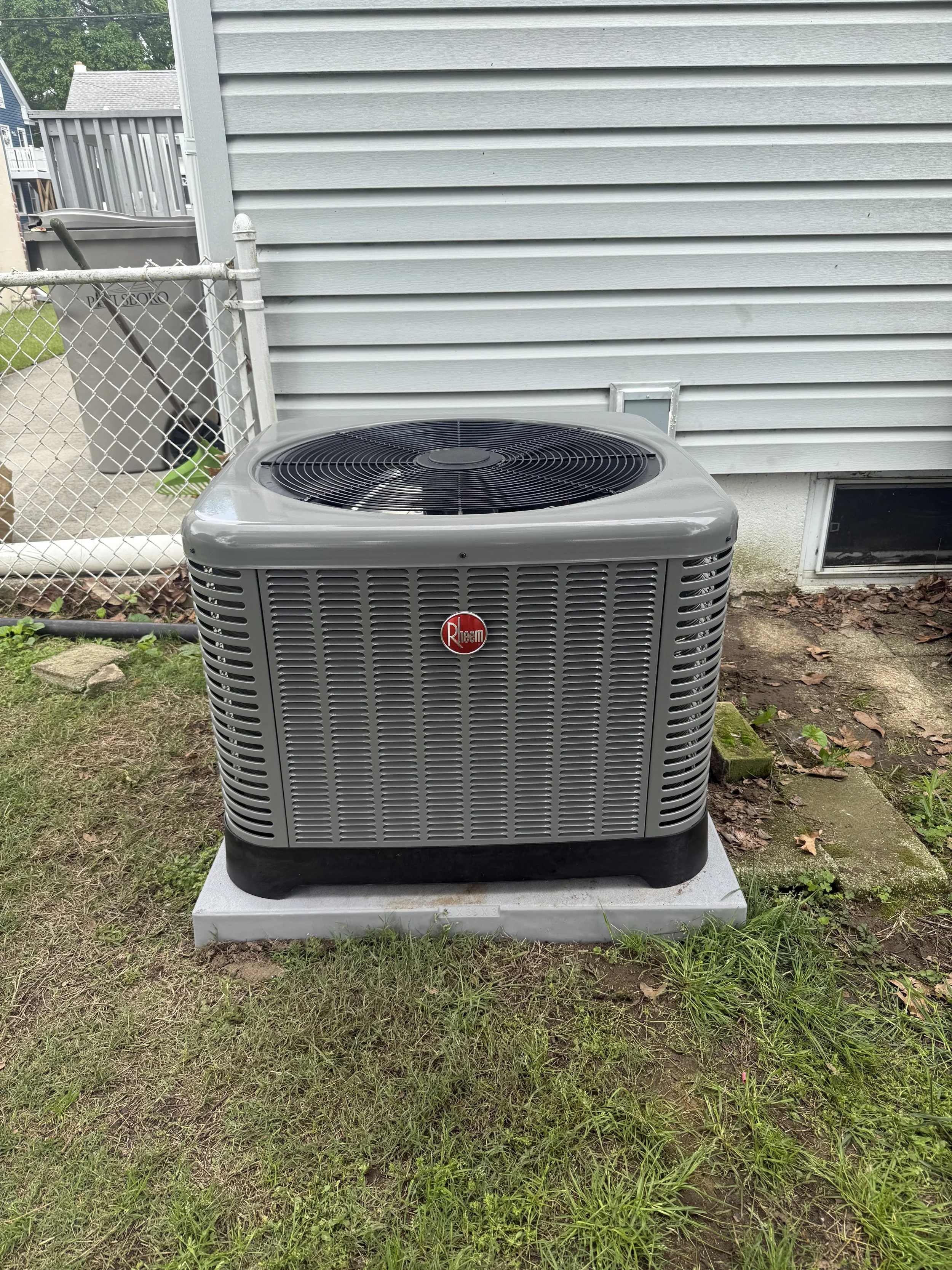 An outdoor Rheem air conditioning unit installed on a concrete pad next to a house with grey vinyl siding, a chain-link fence, and a trash bin in the background.
