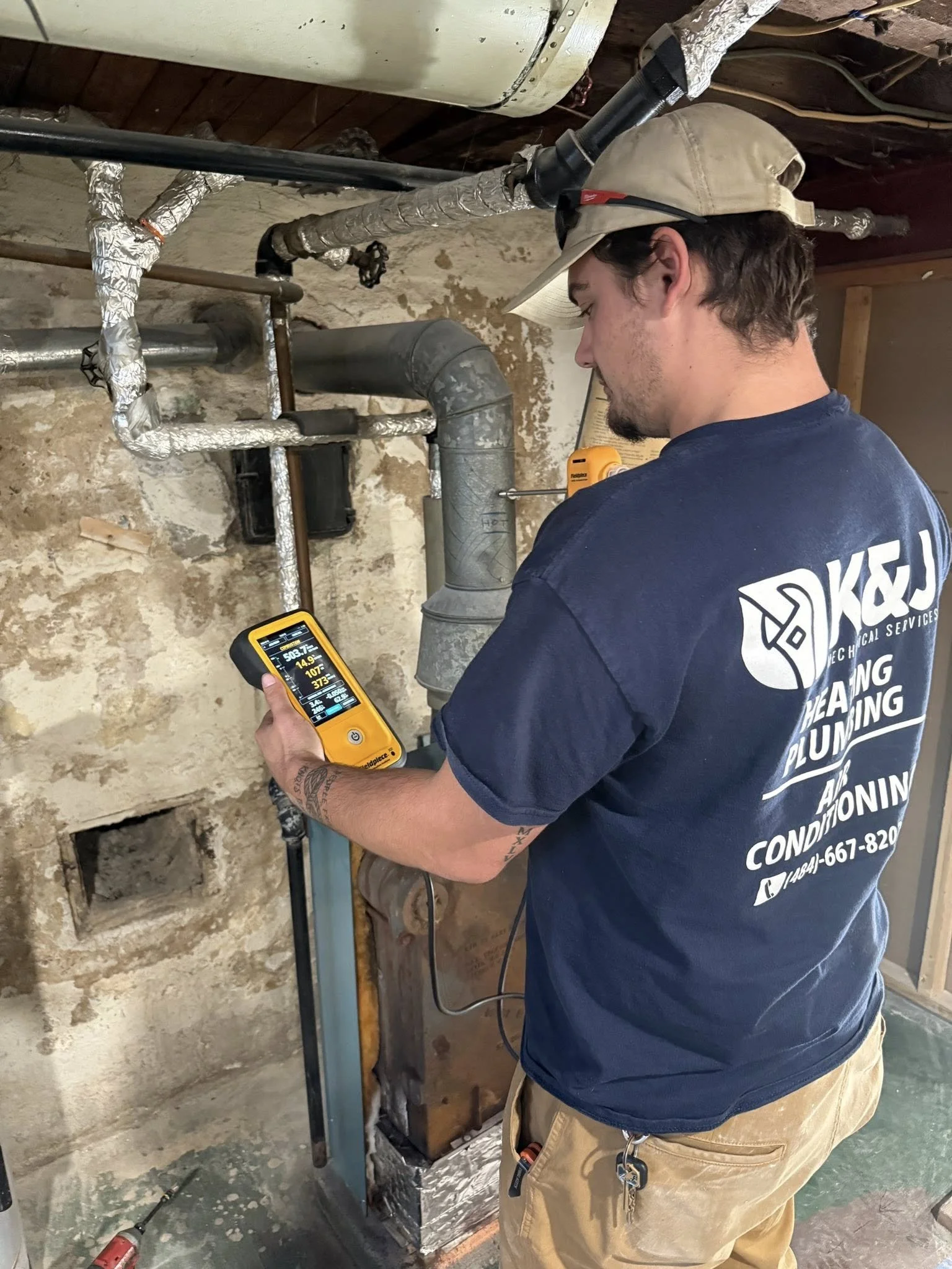 A plumber wearing a navy blue shirt, khaki pants, and a beige cap is inspecting pipework in a basement with a thermal imaging device. The basement wall is old and worn with exposed pipes wrapped in insulation.