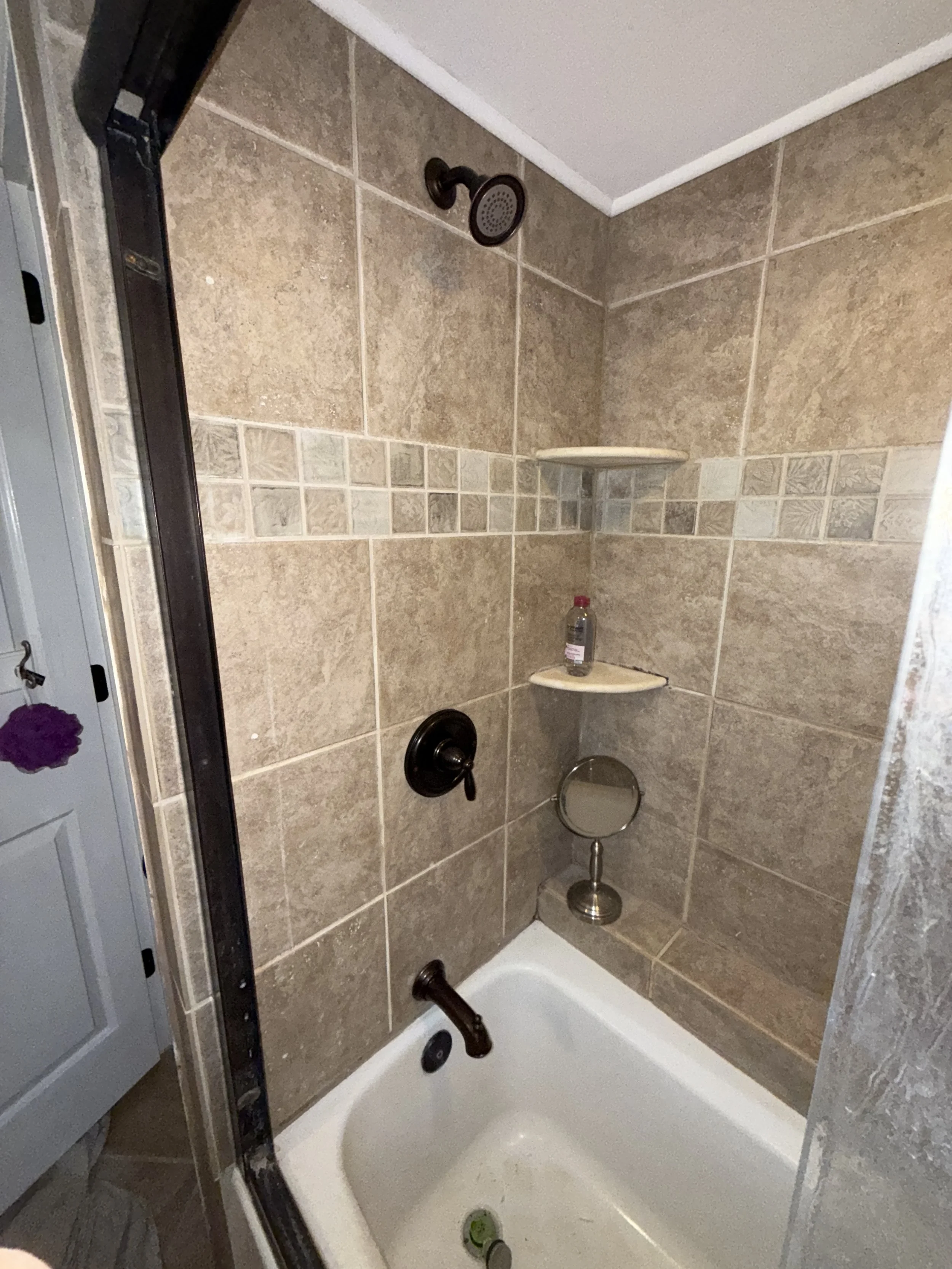 A shower stall with beige tile walls, a dark showerhead, a black faucet, and two corner shelves. A bottle of soap is on the lower shelf, and a small mirror is attached to the wall at the lower right. The shower floor is white with a drain, and a smal