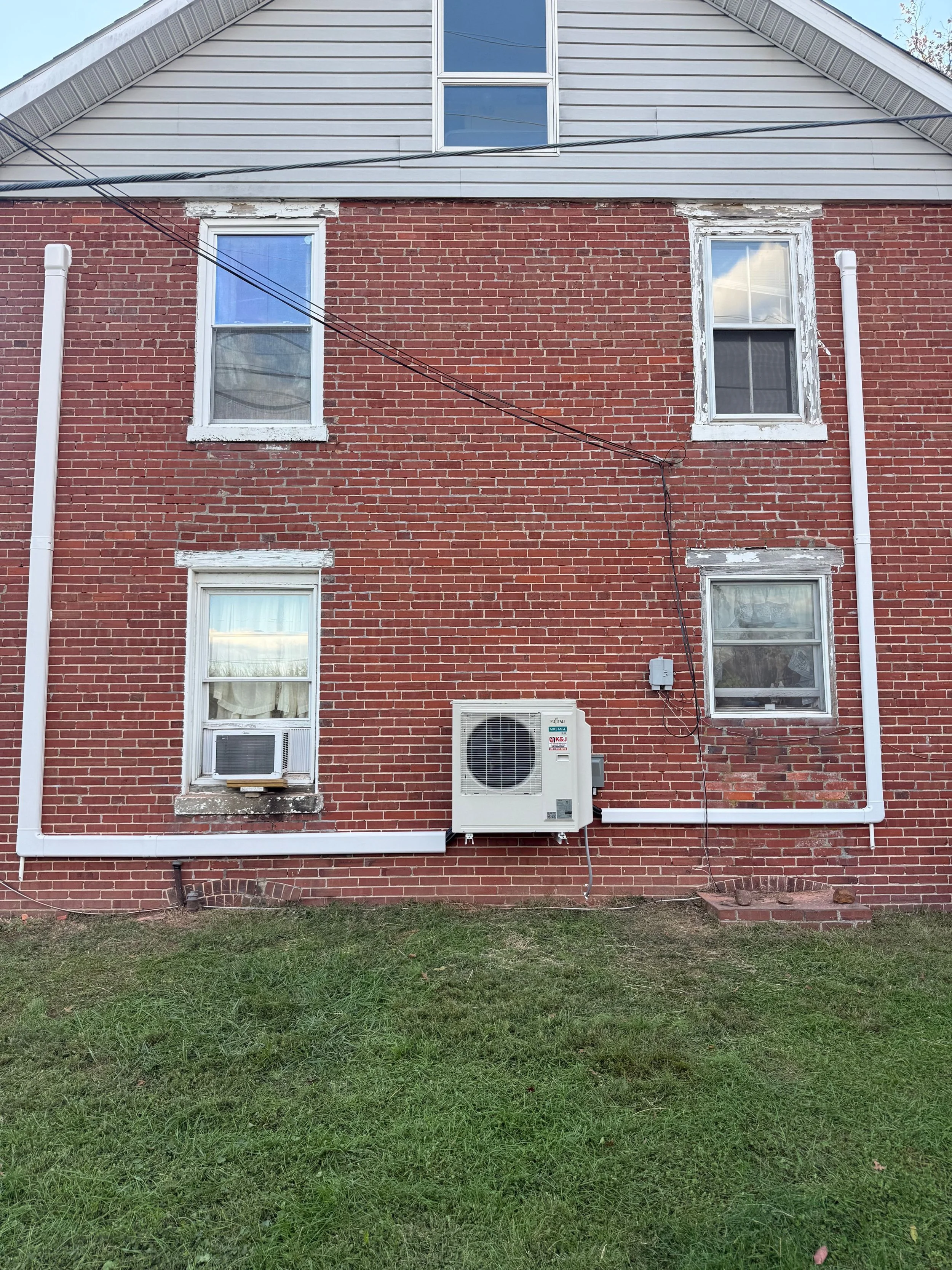 Back of a brick house with four windows, two on the upper floor and two on the lower, air conditioning unit, and white PVC pipes. The house has a gray siding section on the top and a lawn in the foreground.