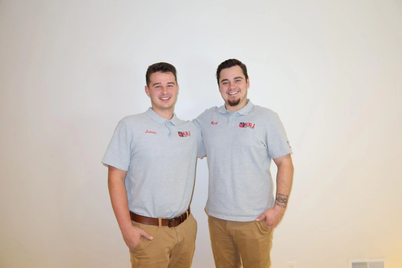 Two young men standing close together, smiling at the camera, against a plain white wall. They are wearing matching light gray polo shirts with red embroidery and tan pants.