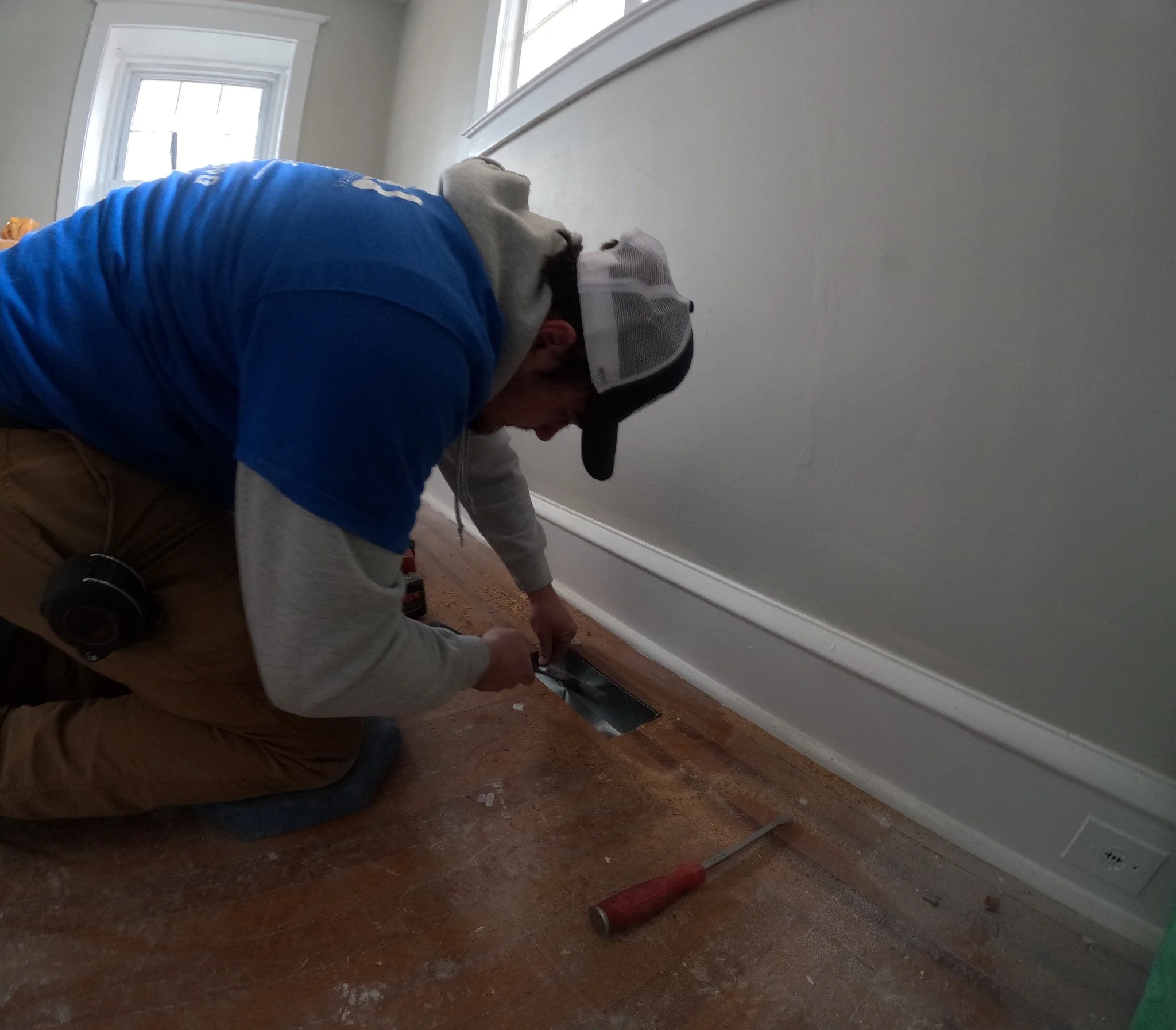 A person kneeling on the floor, working on a vent in a home, wearing a blue hoodie, brown pants, a gray hoodie underneath, and a baseball cap, with a screwdriver nearby.