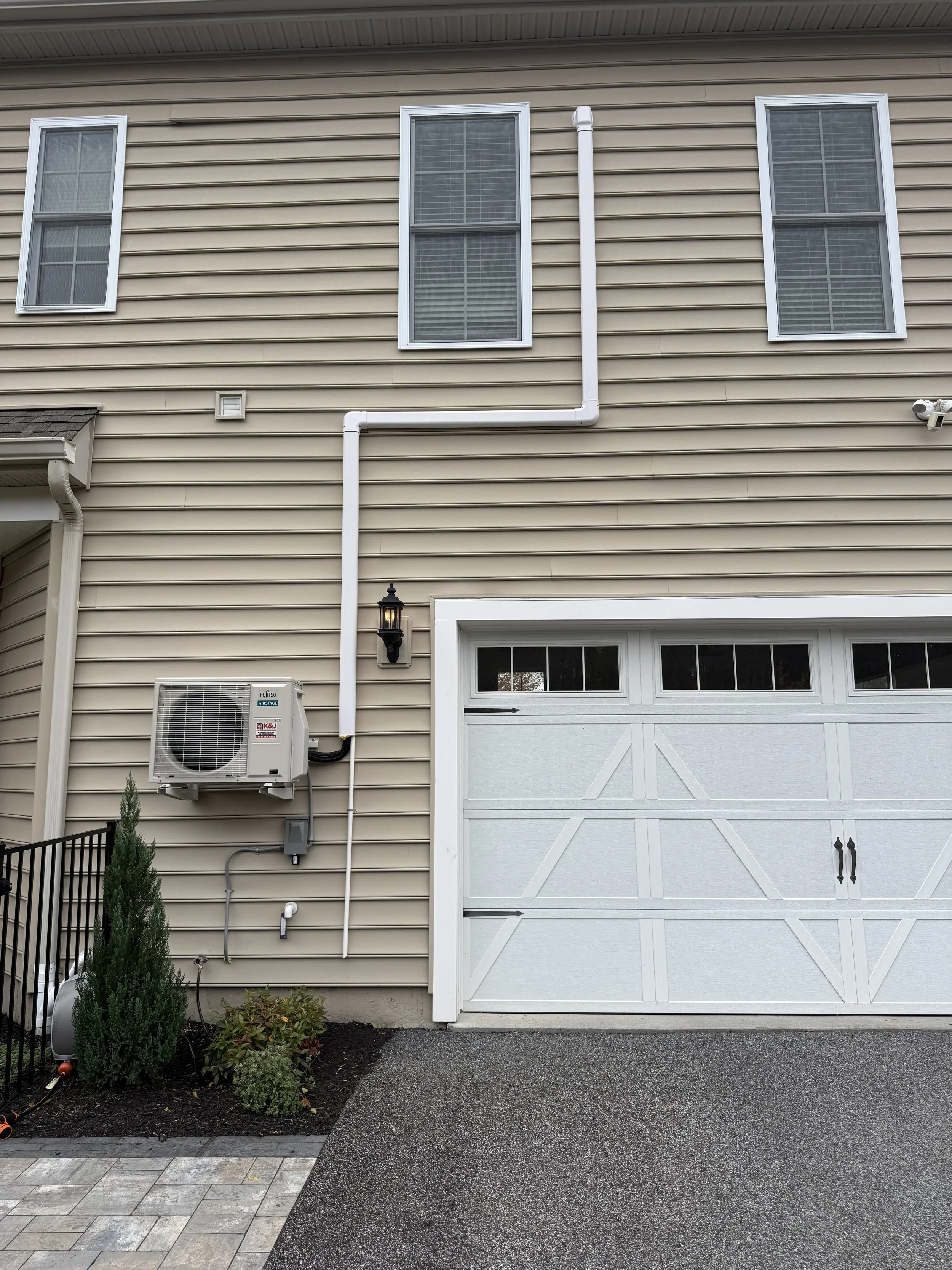 The exterior of a beige house with three tall windows, white trim, a white garage door, an outdoor light fixture, and a utility unit on the wall. There is a small landscaped area with a shrub and plants, and part of a paved walkway.