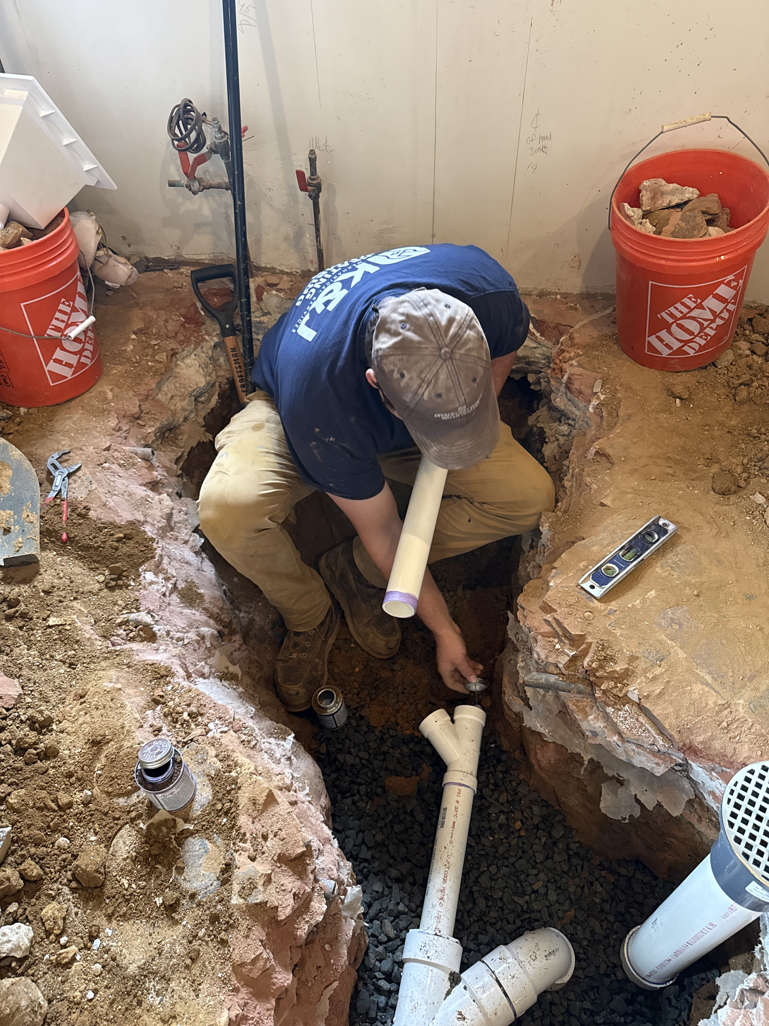 A construction worker installing plumbing pipe in a hole dug in the floor of a building under renovation, with buckets of rocks and construction tools around.