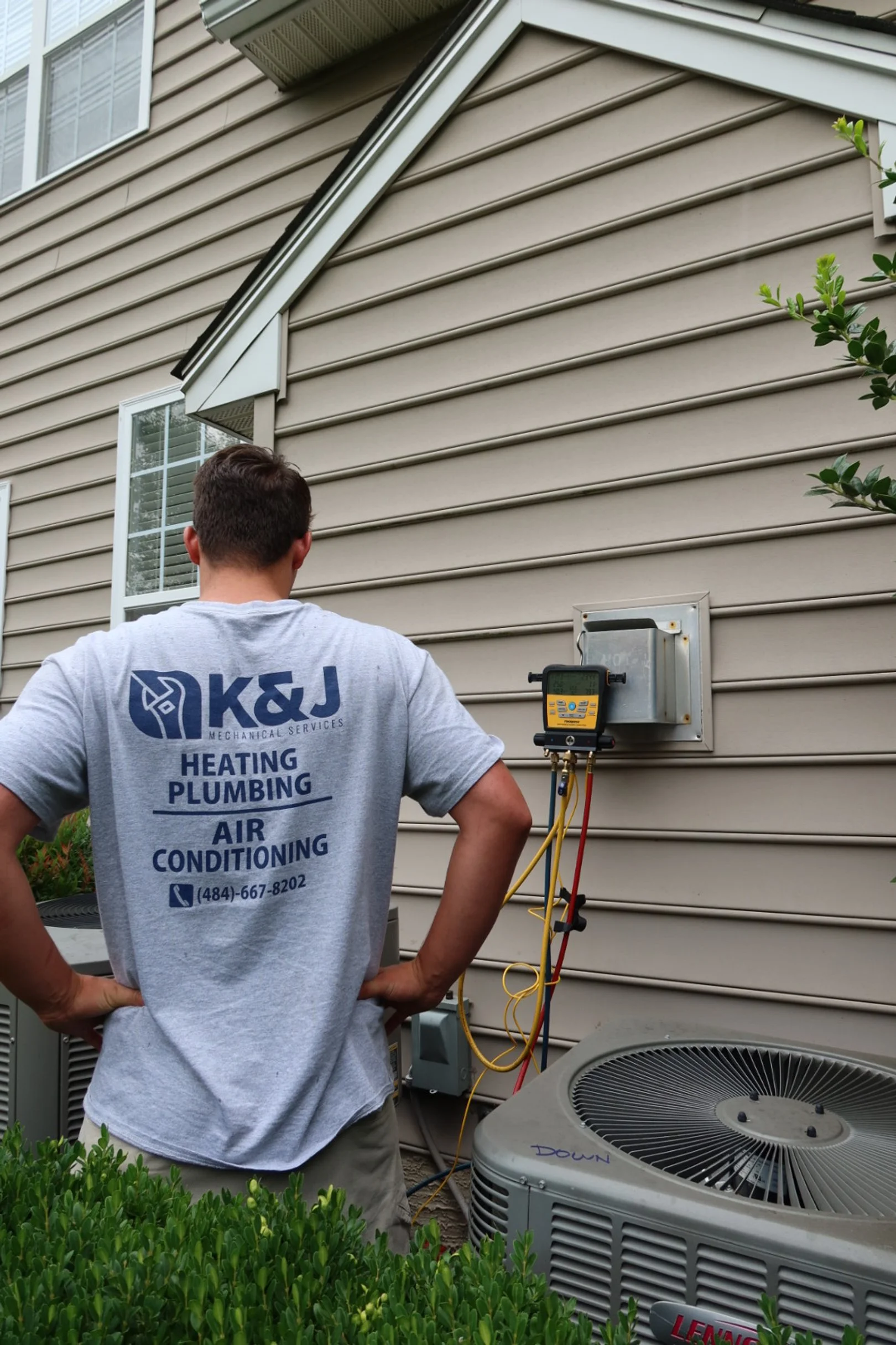 A technician from K&J Mechanical Services working on an air conditioning unit outside a house, with wiring and tools attached to the exterior wall.
