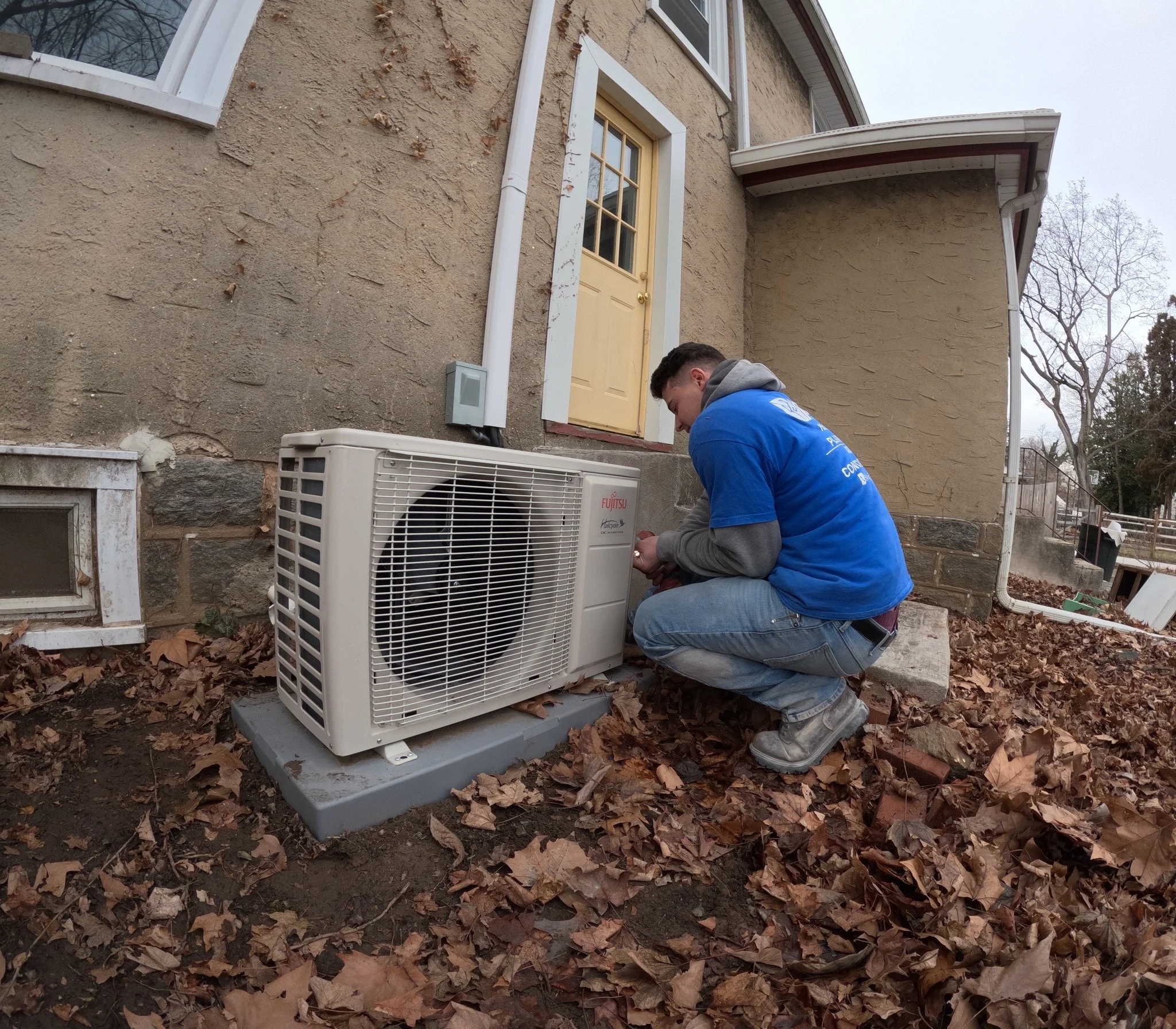 A technician crouched in front of a window-mounted air conditioning unit on the ground outside a house, working on the unit on a fall day with fallen leaves around.