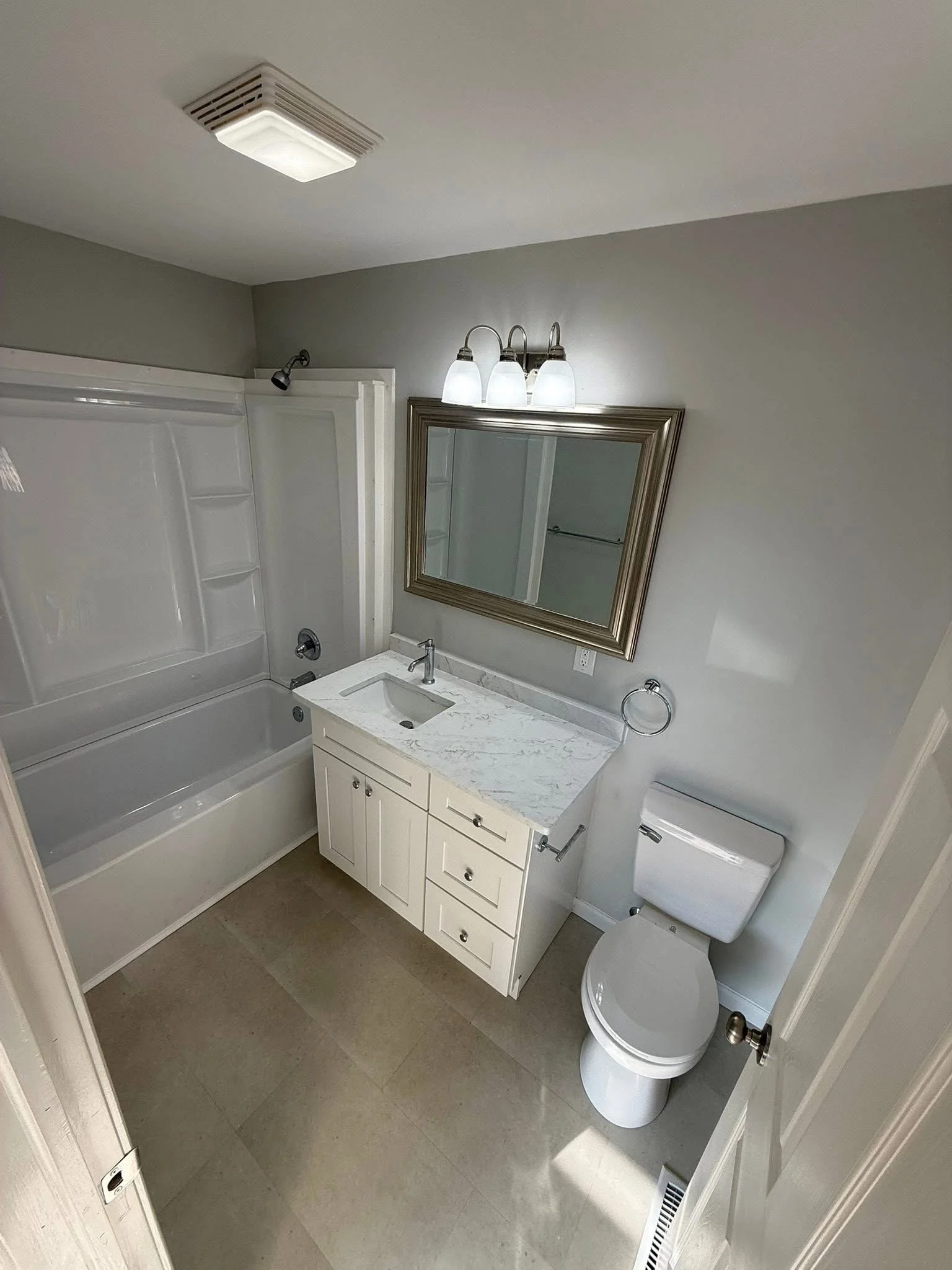 A small bathroom with a bathtub and shower, white vanity with a marble countertop, mirror with a silver frame, three-light fixture, toilet, and tiled floor.