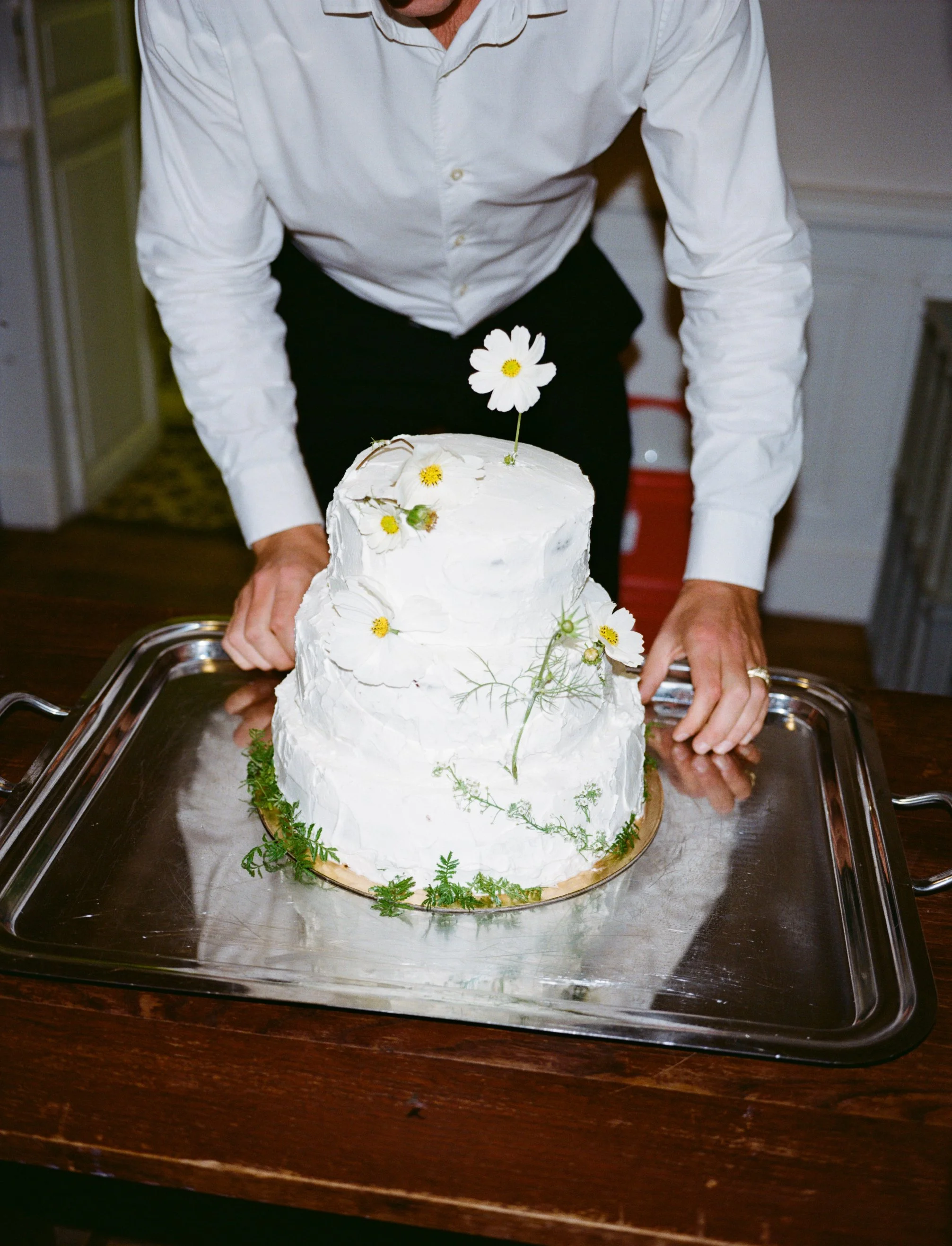 A person in a white shirt is holding a two-tiered white cake decorated with daisies and greenery, placed on a silver tray and a wooden table.