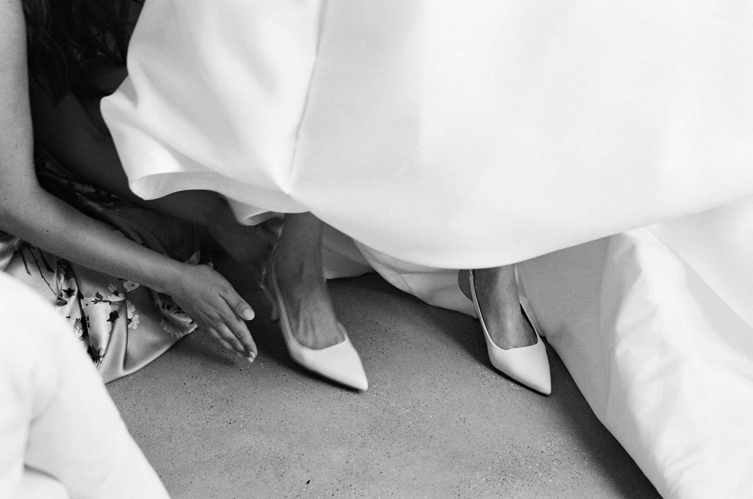 Close-up of women in white high heels, with one woman’s hand resting on her ankle, partially hidden under a white fabric, in black and white.