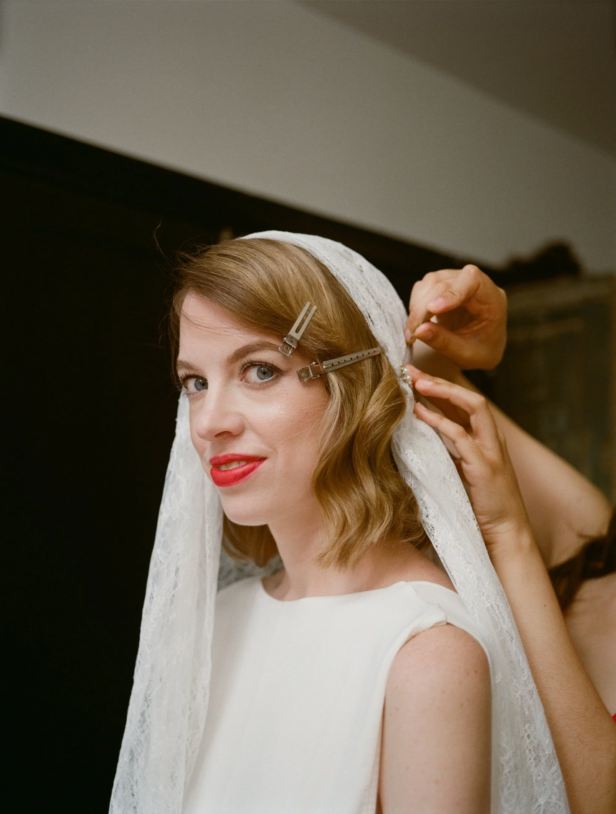 Bride with light brown hair, blue eyes, wearing a white dress and a lace veil, getting her hair styled with hair clips by a stylist.