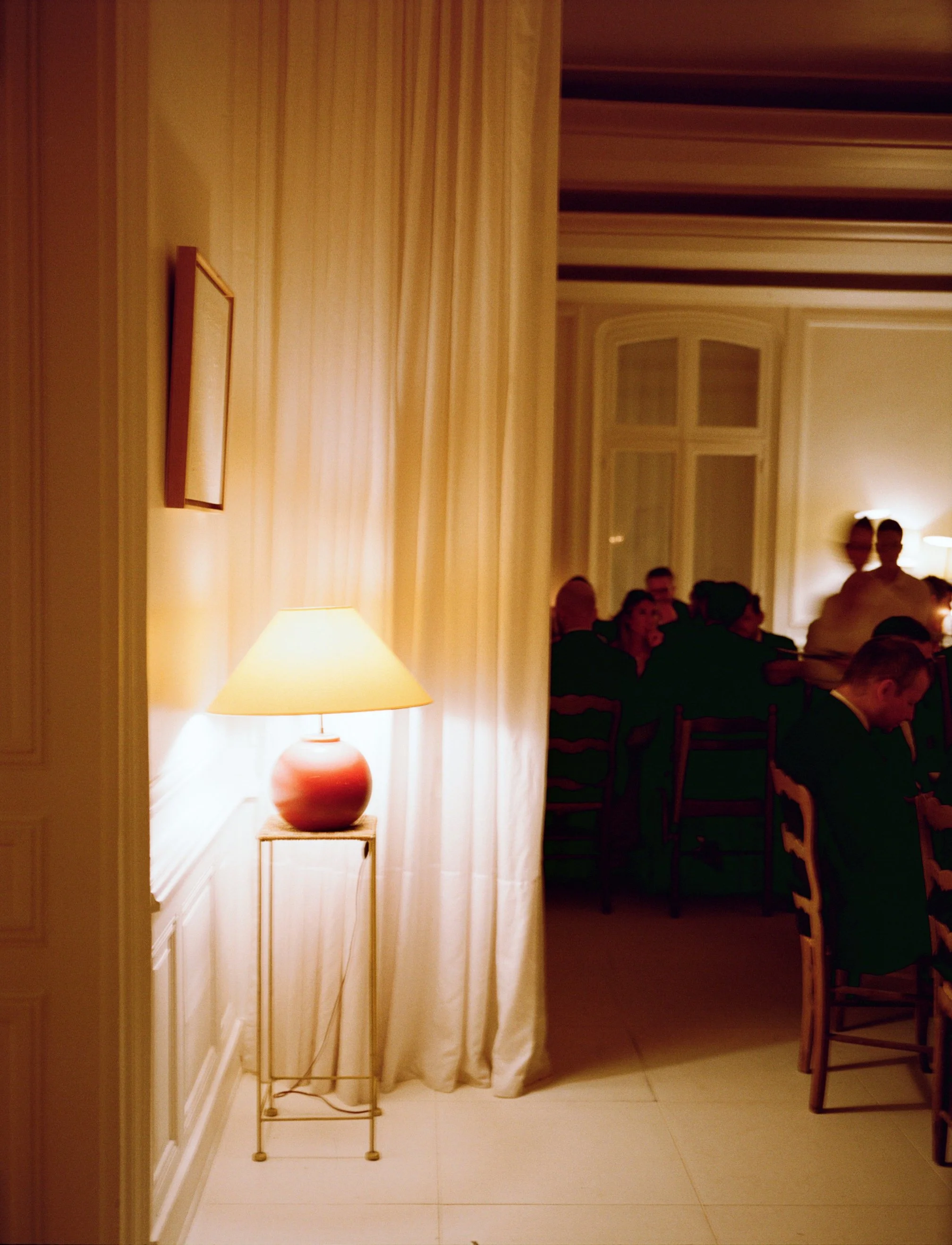 A warmly lit room with beige curtains, a table lamp on a small stand, and a gathering of people seated at a long table in the background.
