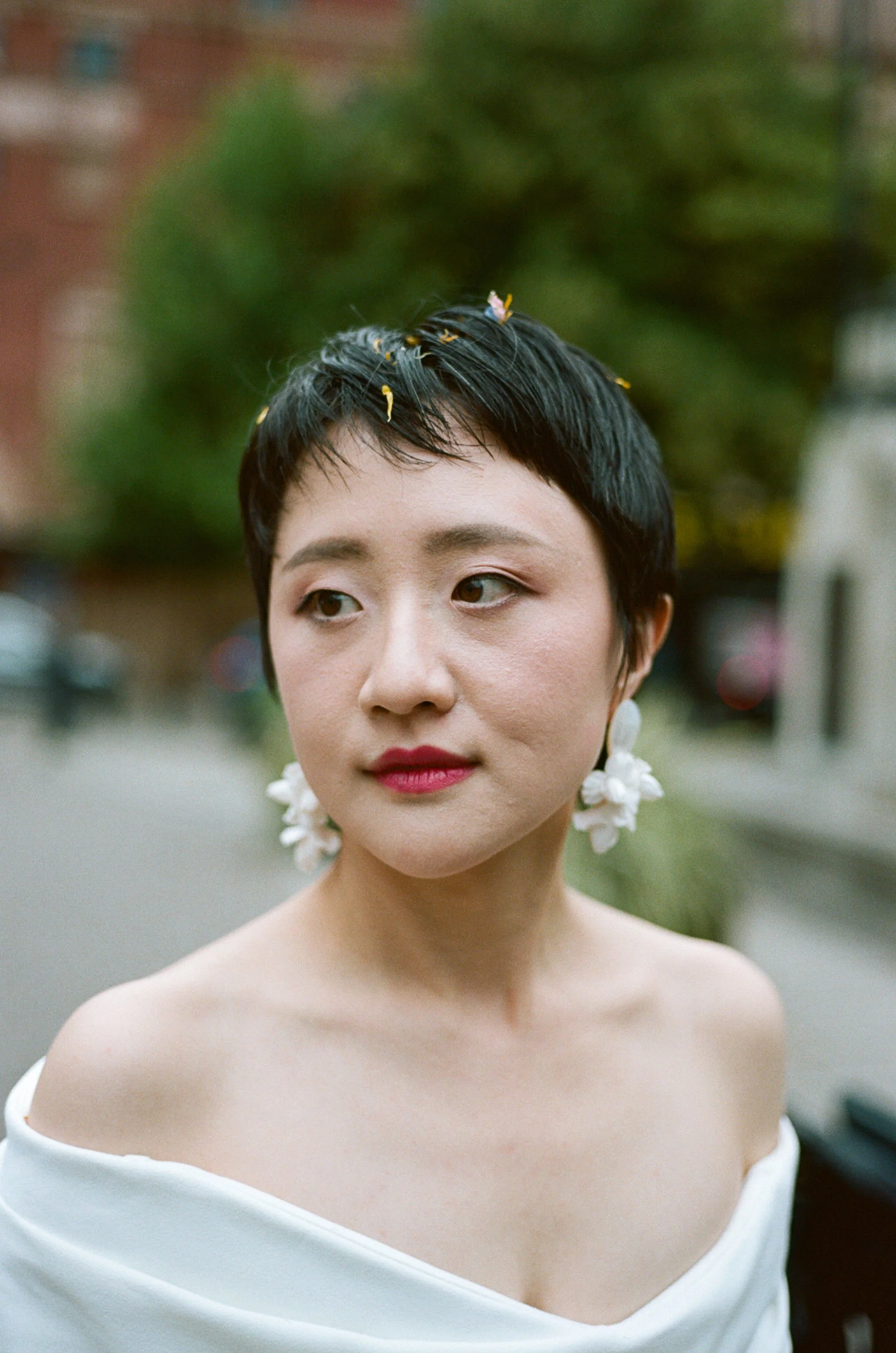Close-up of a woman with short black hair, wearing white floral earrings, an off-shoulder white top, with small flower petals in her hair, standing outdoors with blurred greenery and buildings in the background.