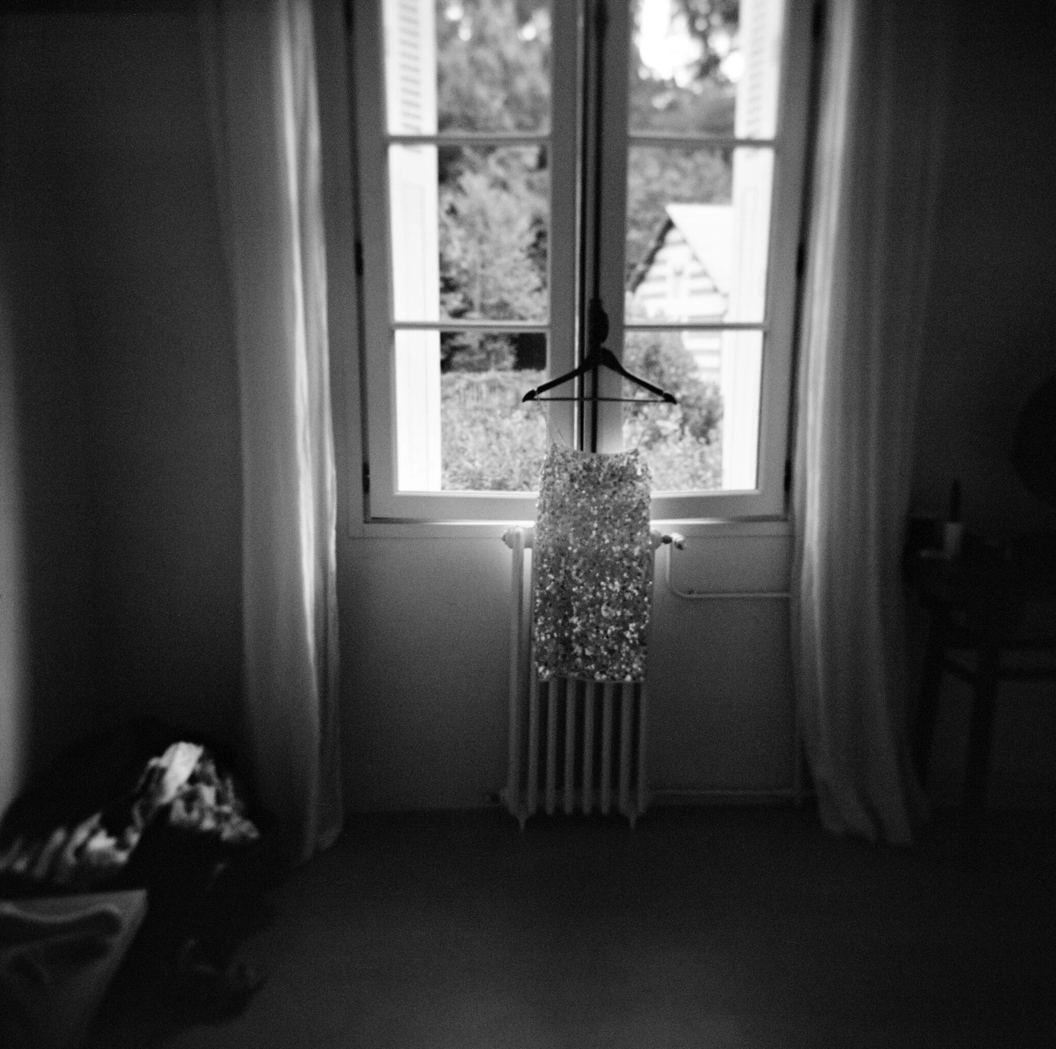 A black and white photo of a bedroom window with a dress hanging on a hanger in front of it. The window has open shutters, and there are trees and a building roof visible outside. The dress appears to be sparkly or sequined.