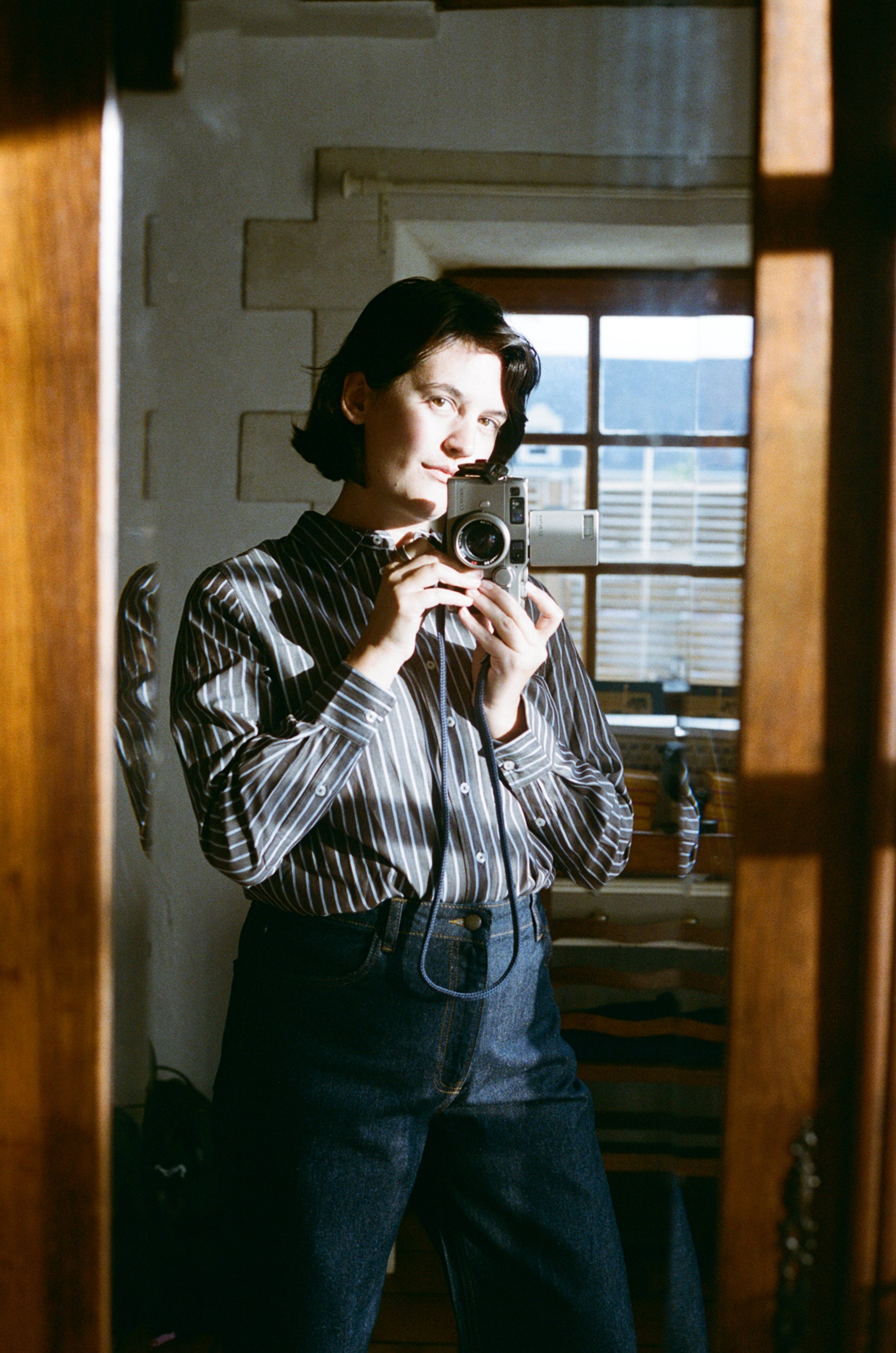 A woman taking a selfie in a mirror with a camera, standing in a room with natural light from a window.
