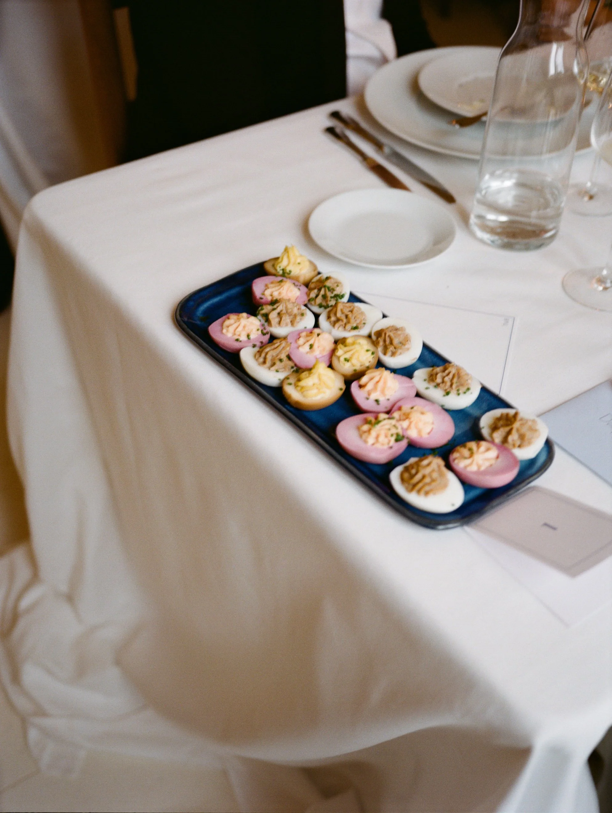 Plate of assorted deviled eggs with pink, yellow, and white toppings on a dark blue platter on a white tablecloth at a formal dining setting.
