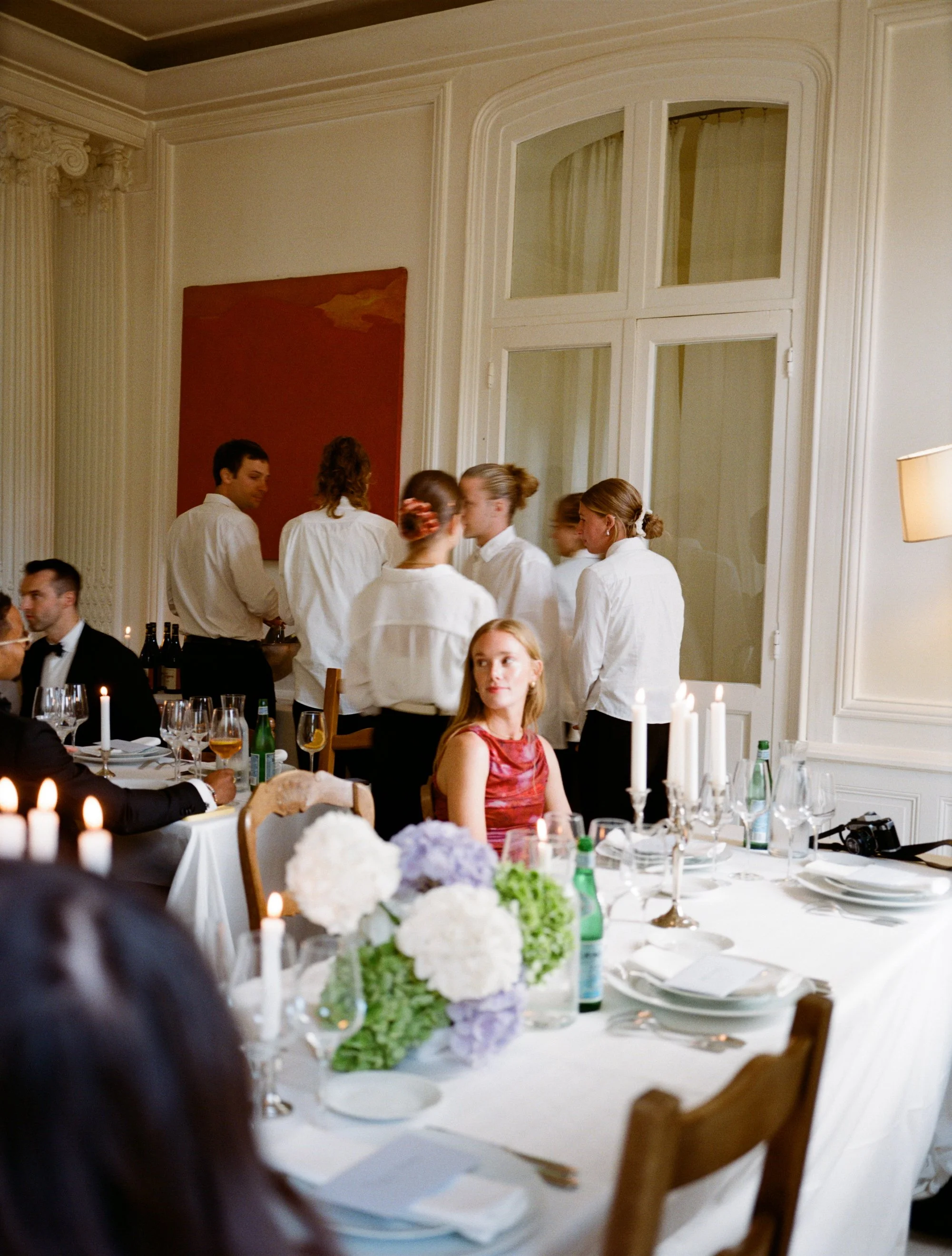 A formal dining room with white tablecloths, floral centerpieces, and multiple servers dressed in white shirts. Some guests are seated, while others are standing near a large window, creating a sophisticated atmosphere.