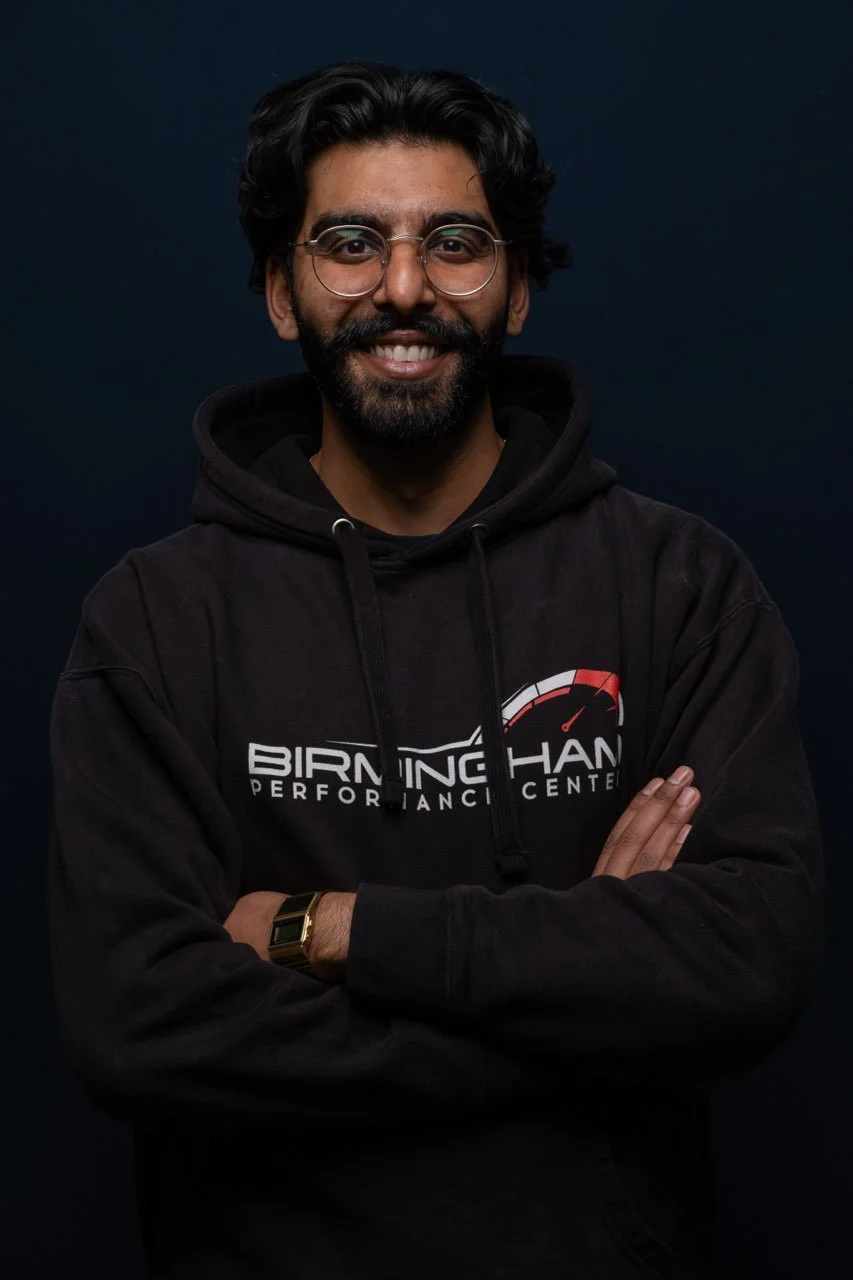 Smiling man with glasses, beard, and crossed arms wearing a black hoodie with a performance center logo, standing against a dark background.
