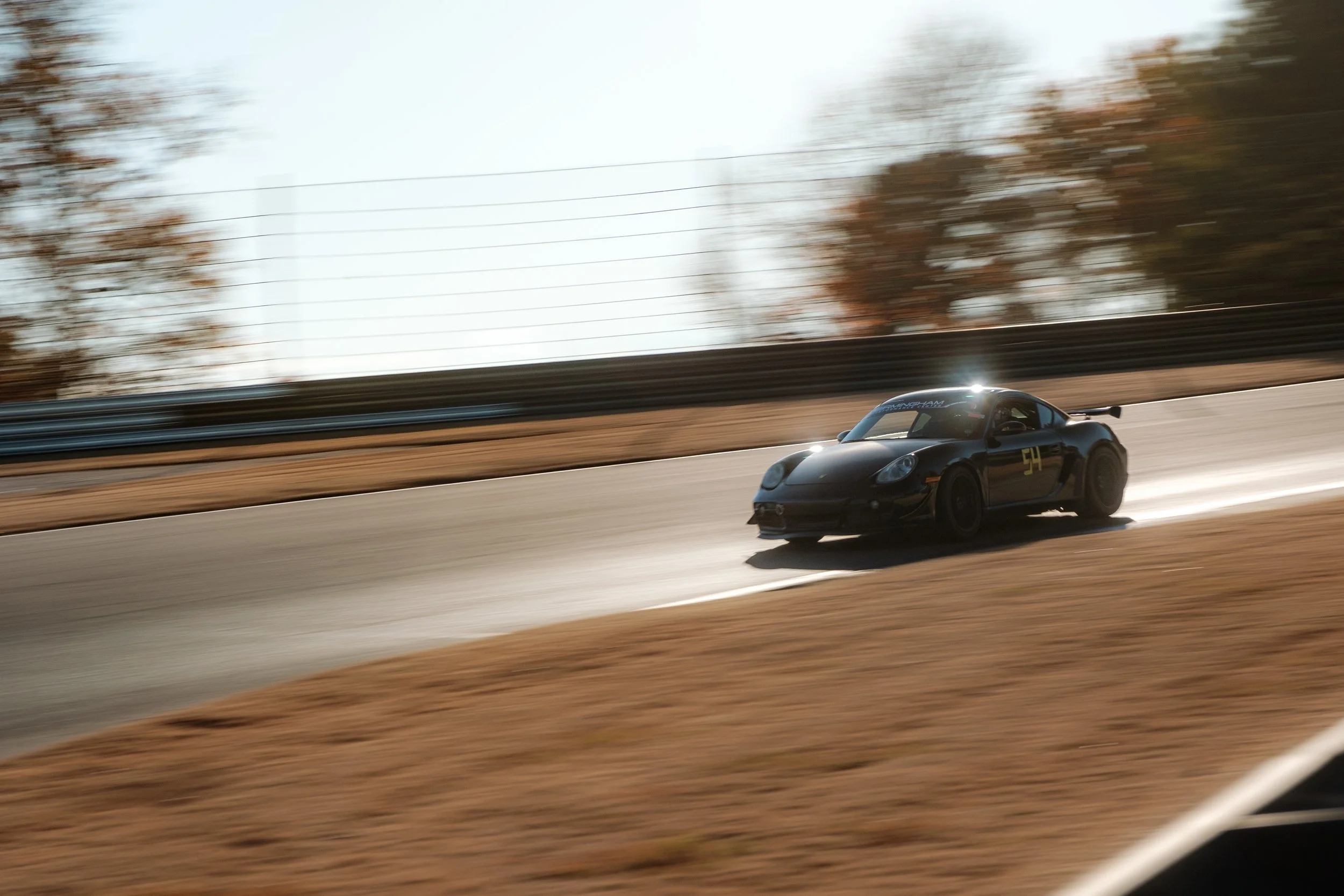 A black sports car racing on a track during daytime with motion blur, trees and a fence in the background.